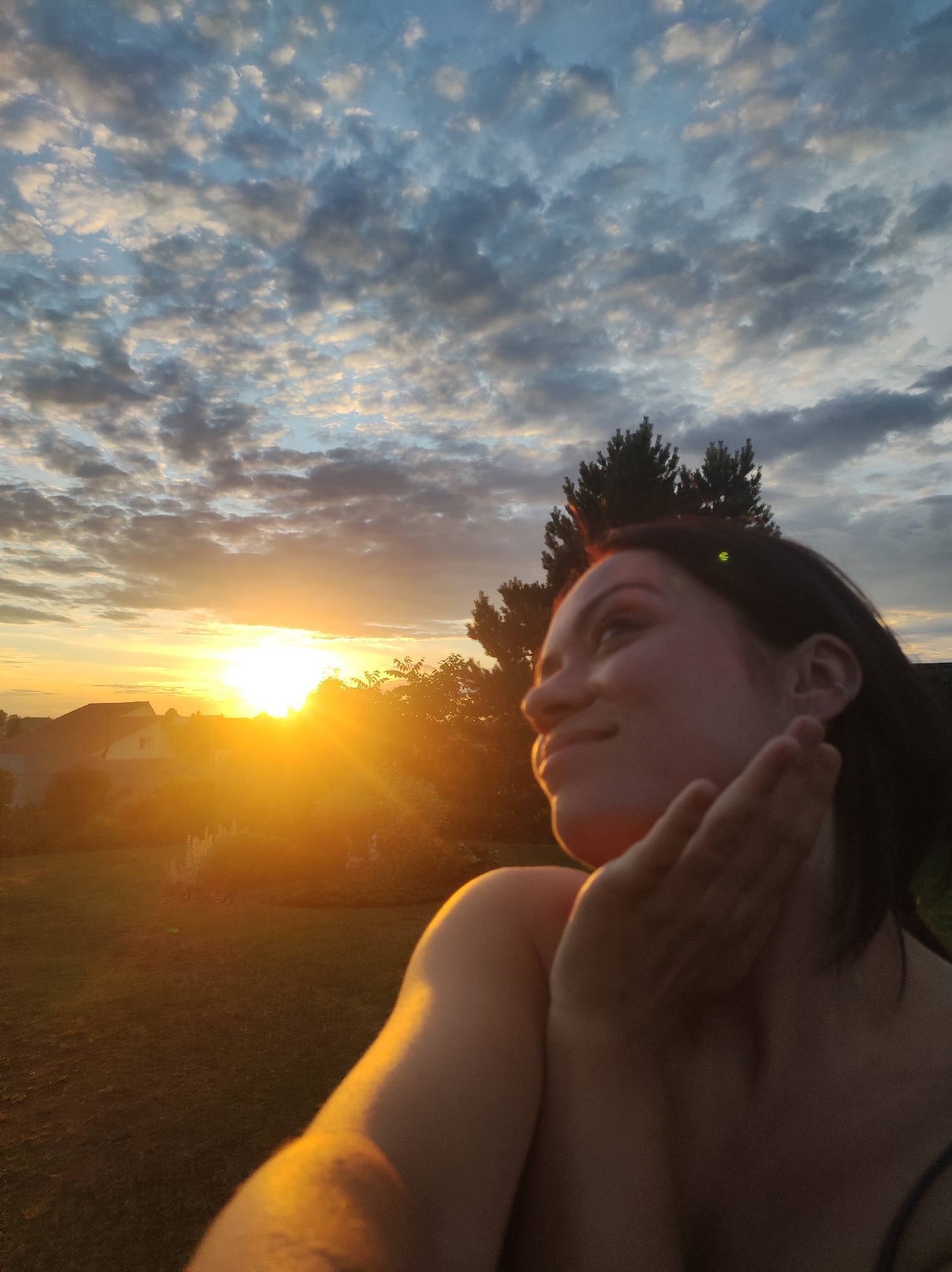 The media file captures a young woman outdoors during a sunset. The sun, a bright orange orb, is low on the horizon, casting a warm golden light across the scene. It is situated in the upper left quadrant of the image, with rays of light emanating outwards, illuminating a distant landscape of houses and trees. The sky above is a mix of blue and scattered clouds, tinged with pink and orange from the setting sun.
The woman is positioned in the right foreground, her face turned upwards and slightly to the left, gazing towards the sunset. Her expression appears serene and contemplative. She is holding one hand up to her face, her fingers softly touching her cheek and chin, partially obscuring her features but adding a sense of intimacy to the shot. Her shoulder and arm are visible in the lower right, bathed in the golden light of the sunset, highlighting her skin. The background behind her includes dark silhouettes of trees, including a tall evergreen.
The setting appears to be a rural or suburban area, suggested by the houses visible in the distance. The ground in the foreground is covered with grass, also illuminated by the warm sunset hues. The time of day is clearly evening, as indicated by the low sun and the colors in the sky. The weather appears clear with scattered clouds, creating a picturesque sunset. The overall mood is peaceful and reflective. There is no visible text in the image. The location context is identified as Фоки, Russia.