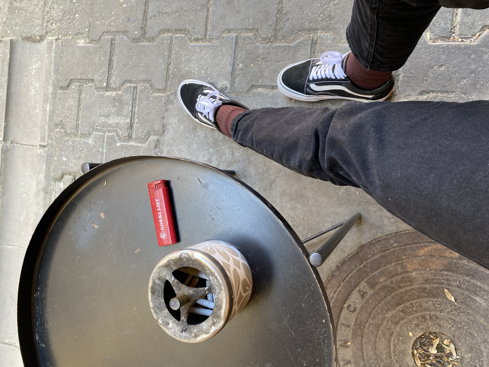 The image shows a partially obscured view of a person seated at a table, likely outdoors. The person is wearing black jeans and black Vans Old Skool sneakers with white laces and brown socks. The person's legs are positioned at an angle across the frame.
A small, round black metal table is visible in the foreground. On the table sits a red lighter with the text "GORKI LIST" printed on it. Next to the lighter is a ceramic ashtray with a decorative leaf pattern. The ashtray is filled with cigarette butts.
The table is situated on a paved surface made of interlocking gray bricks. A circular metal manhole cover is partially visible to the right of the table, near the person's leg. The manhole cover has concentric circle patterns and inscriptions.
The location is identified as Novi Sad, Serbia, based on the "Gorki List" lighter, which is a product commonly associated with Serbia and the surrounding region.