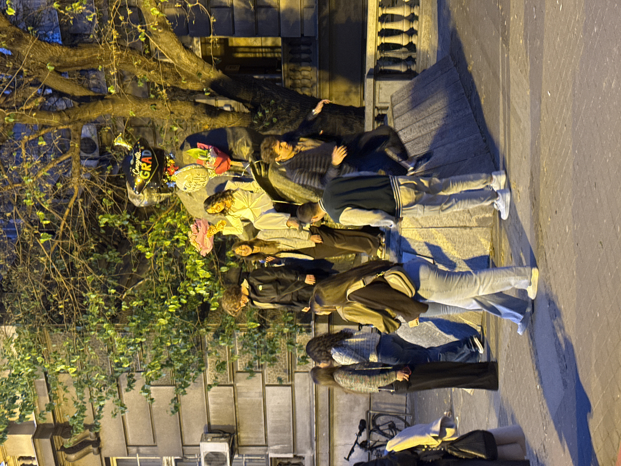 A group of people are gathered in Belgrade, Serbia, sitting and lying near a tree. The event appears celebratory, with graduation balloons and flowers present. People are dressed casually. A bicycle is visible near the edge of the sidewalk, and the street is lit. The architecture of the building in the background shows ornate detailing.