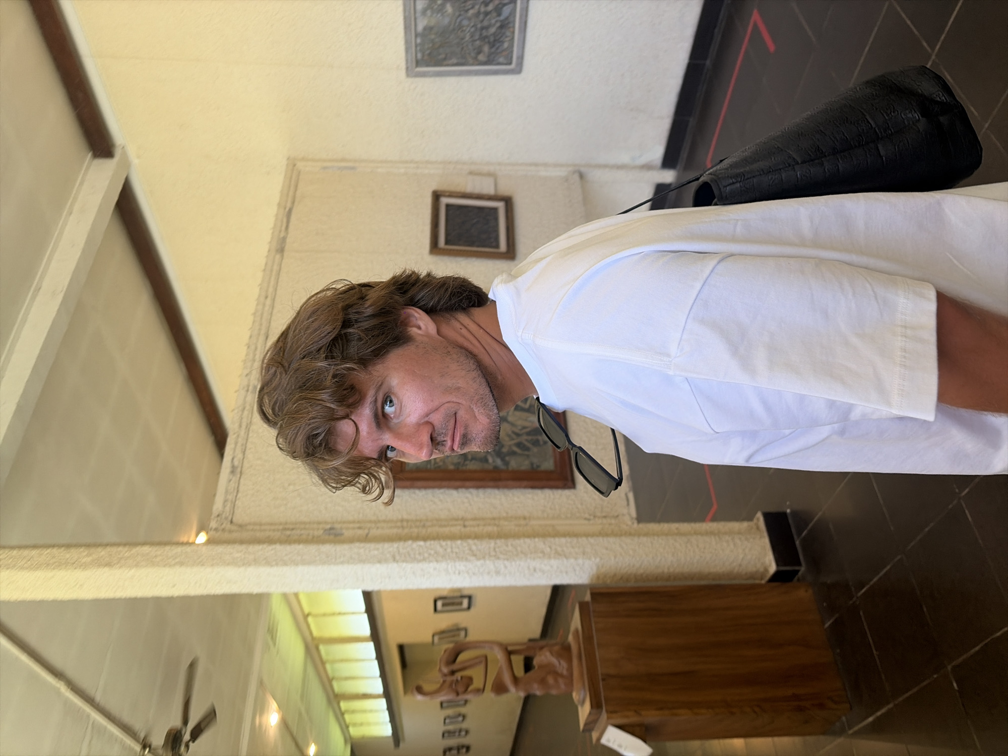 A man with tousled brown hair and a light beard is looking over his shoulder at the camera in a museum in Ubud, Indonesia. He is wearing a white t-shirt with sunglasses hanging from his neck and a black leather purse slung over his shoulder. The man is standing in a gallery with a polished dark floor and white walls. To his left, there is a wooden pedestal with a wooden sculpture on it, and a white sign. Further down the gallery, there are several framed artworks on the wall. The ceiling is high with a fan and recessed lighting. The man appears to be posing for a photograph.