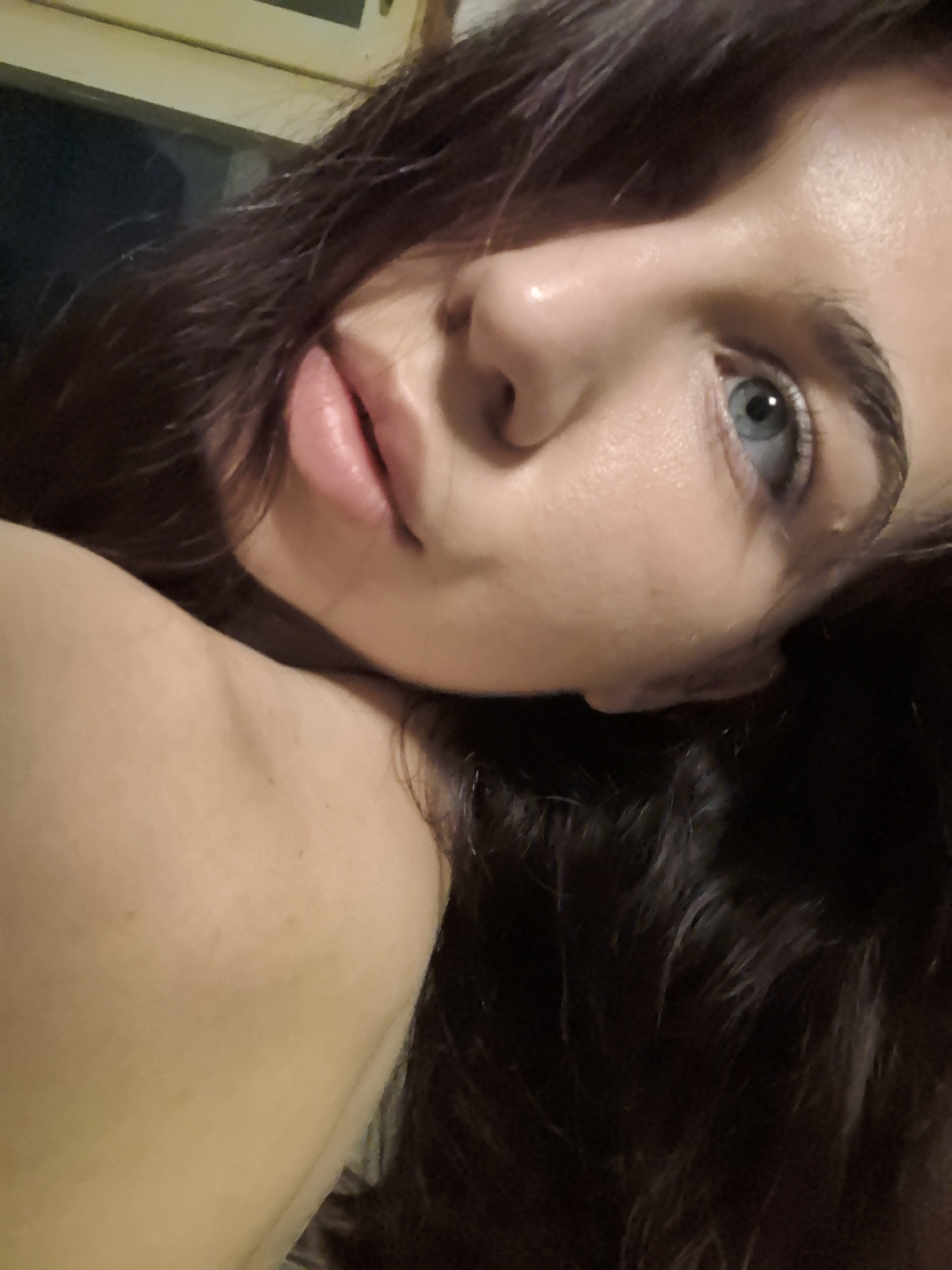 A close-up shot of a woman with dark hair and light blue eyes, likely taken indoors. Her skin is fair, and her lips are a soft pink. The background is dark and indistinct. Her shoulder is visible. The location is Unknown, Unknown.