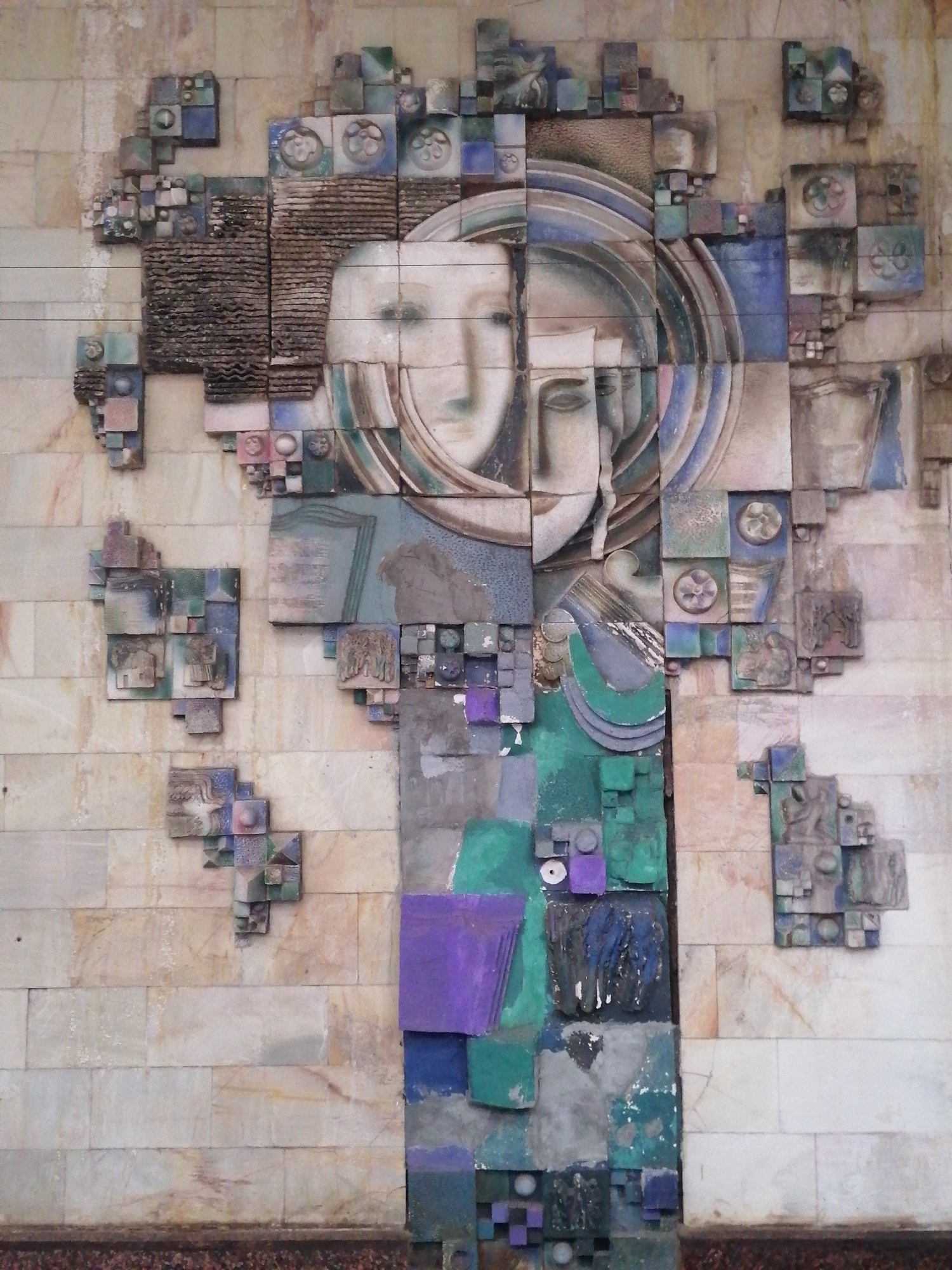 This is a photograph of a large, intricate, relief sculpture adorning a building facade. The sculpture is made of various materials, including what appears to be concrete, ceramic tiles, and possibly metal, in a palette of muted earth tones, blues, greens, and purples. The main subject of the sculpture is a stylized, almost abstract depiction of two faces, rendered in profile and overlapping. The faces are pale and serene, with large, almond-shaped eyes.
The sculpture is highly textured and geometric, incorporating numerous cubical and rectangular elements, as well as circular motifs and abstract shapes. Some of these elements are arranged in clusters resembling architectural structures or intricate machinery. Small, embossed or carved details are visible on some of the square tiles, including what seem to be floral patterns and miniature scenes or figures.
The sculpture is mounted on a wall constructed of large, rectangular, light-colored stone tiles, possibly marble or granite. The overall impression is one of artistic abstraction and complexity, with the faces forming a central, human element within a more abstract, fragmented composition. There is no visible text in the image. The lighting suggests it was taken during daylight. Given the context, this is likely a public art installation in Tashkent, Uzbekistan. The artwork evokes a sense of introspection and perhaps a commentary on the duality of human experience or perception.