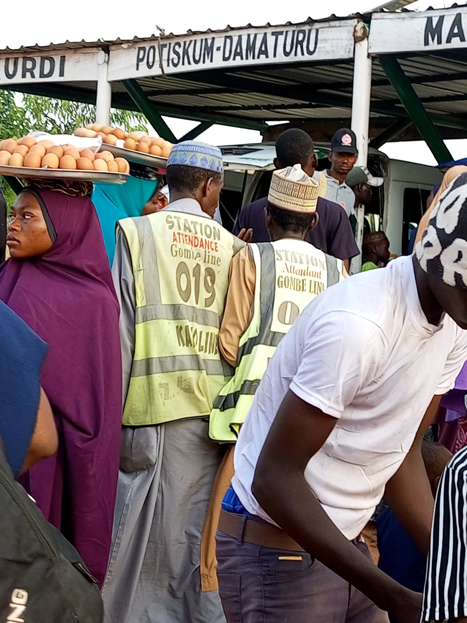 A medium shot captures an outdoor scene at a bus station in Gombe, Nigeria. A woman wearing a purple hijab and a dark headscarf stands to the left, her face turned to the left with a pensive expression. She has a tray of eggs balanced on her head. To her right, a man in a yellow vest with "STATION ATTENDANCE GOMBE LINE" and "019" printed on the back is facing away from the camera. He wears a white cap with blue and green embroidery. Behind him, a man in a grey shirt and a black baseball cap with a logo is looking to his right. Further in the background, a white bus is visible. The station building has a white roof and a sign that reads "KURDI POTISKUM-DAMATURU MA". The ground appears to be dirt.