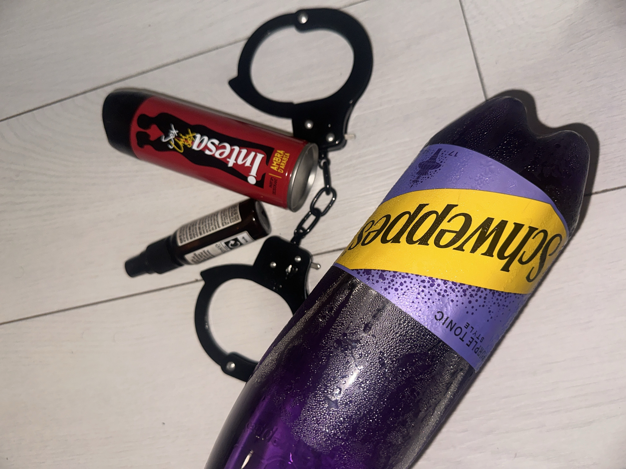 The scene depicts four distinct objects arranged on a light-colored, planked wooden floor. A 1.5-liter plastic bottle of Schweppes Purple Tonic lies diagonally across the lower left and center of the frame, containing a purple liquid and covered in condensation, indicating a low temperature. The bottle's label features prominent yellow "Schweppes" branding and a purple band with "PURPLE TONIC STYLE" printed on it. To the right of the Schweppes bottle, a pair of black metal handcuffs, connected by a chain, are visible. Situated above and partially resting against one of the handcuff rings is a red aerosol can, identified as "Intesa" brand deodorant, specifically "AMBRA D'ARABIA." This can displays a graphic silhouette of a man and woman embracing, with the word "Sex" in yellow script. Adjacent to the Intesa can, slightly to its left, is a small, dark amber-colored glass spray bottle with a black pump-action nozzle. The wooden floor planks exhibit a consistent light tone and visible seams. The scene is illuminated by an overhead light source, casting subtle shadows. The scene is located in the city of Sofia, Bulgaria.