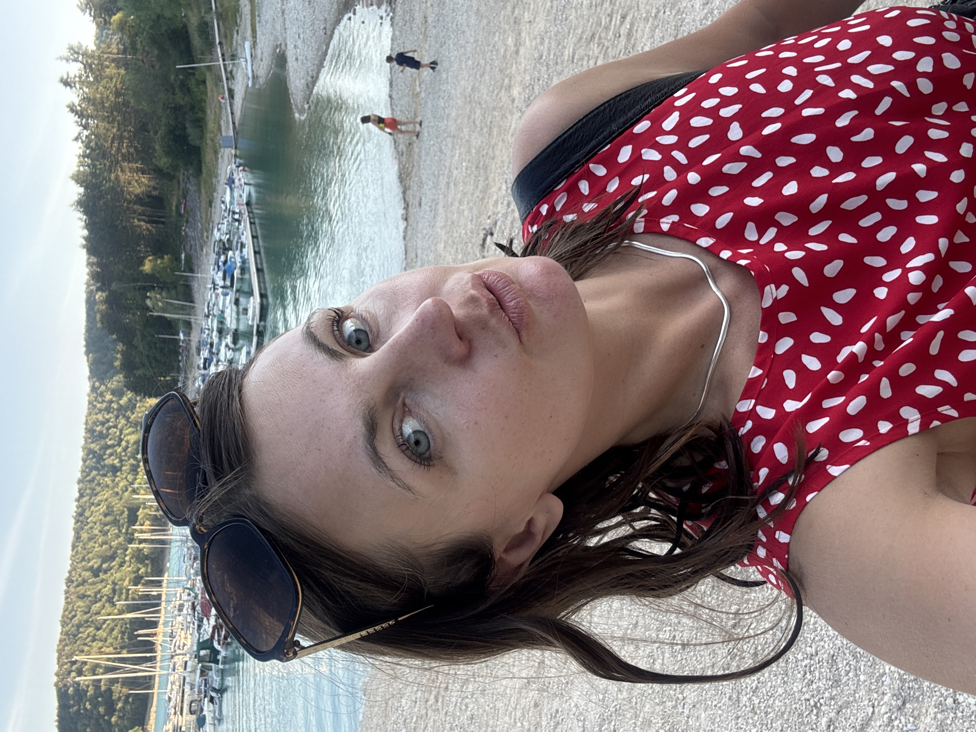 A woman with dark brown hair and light blue eyes, wearing a red dress with white polka dots and a silver necklace, is taking a selfie outdoors. She is positioned in the foreground, looking directly at the camera with her lips pursed. Sunglasses are resting on top of her head. In the background, a body of water with numerous sailboats docked is visible, bordered by a tree-covered hillside. Two children can be seen walking on the pebble beach in the mid-ground. The scene is set at a lake or marina.