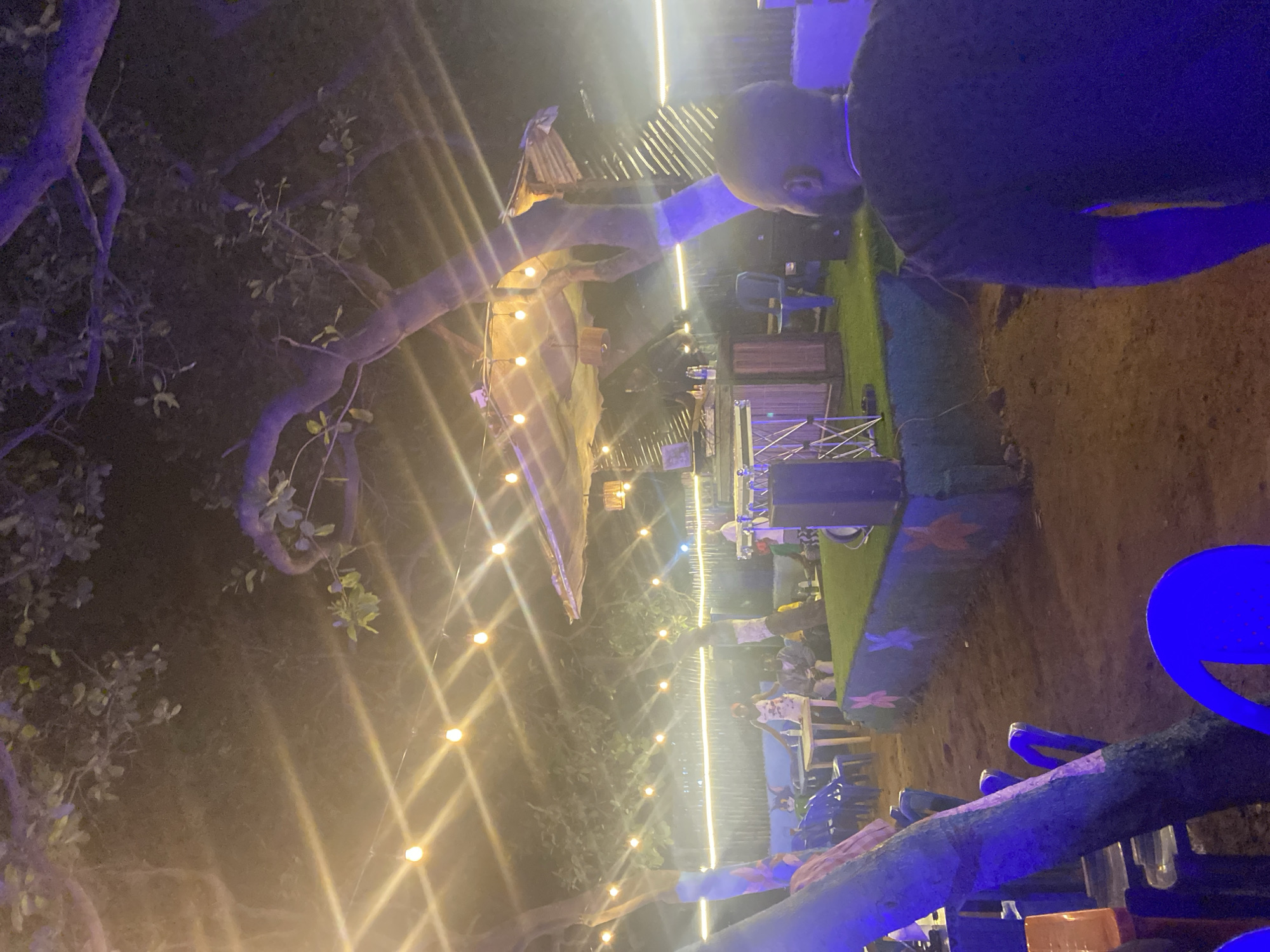 The image depicts an outdoor nighttime scene at a venue in Karum, Nigeria. String lights are strung across the area, illuminating trees and creating a festive atmosphere. In the background, a raised stage is set up with what appears to be DJ equipment, suggesting a musical performance. Several people are visible, some seated at tables and others standing near the stage. The ground is covered in sand, and there are various chairs and tables scattered throughout the venue. The overall impression is of a lively gathering, likely a bar or entertainment spot.