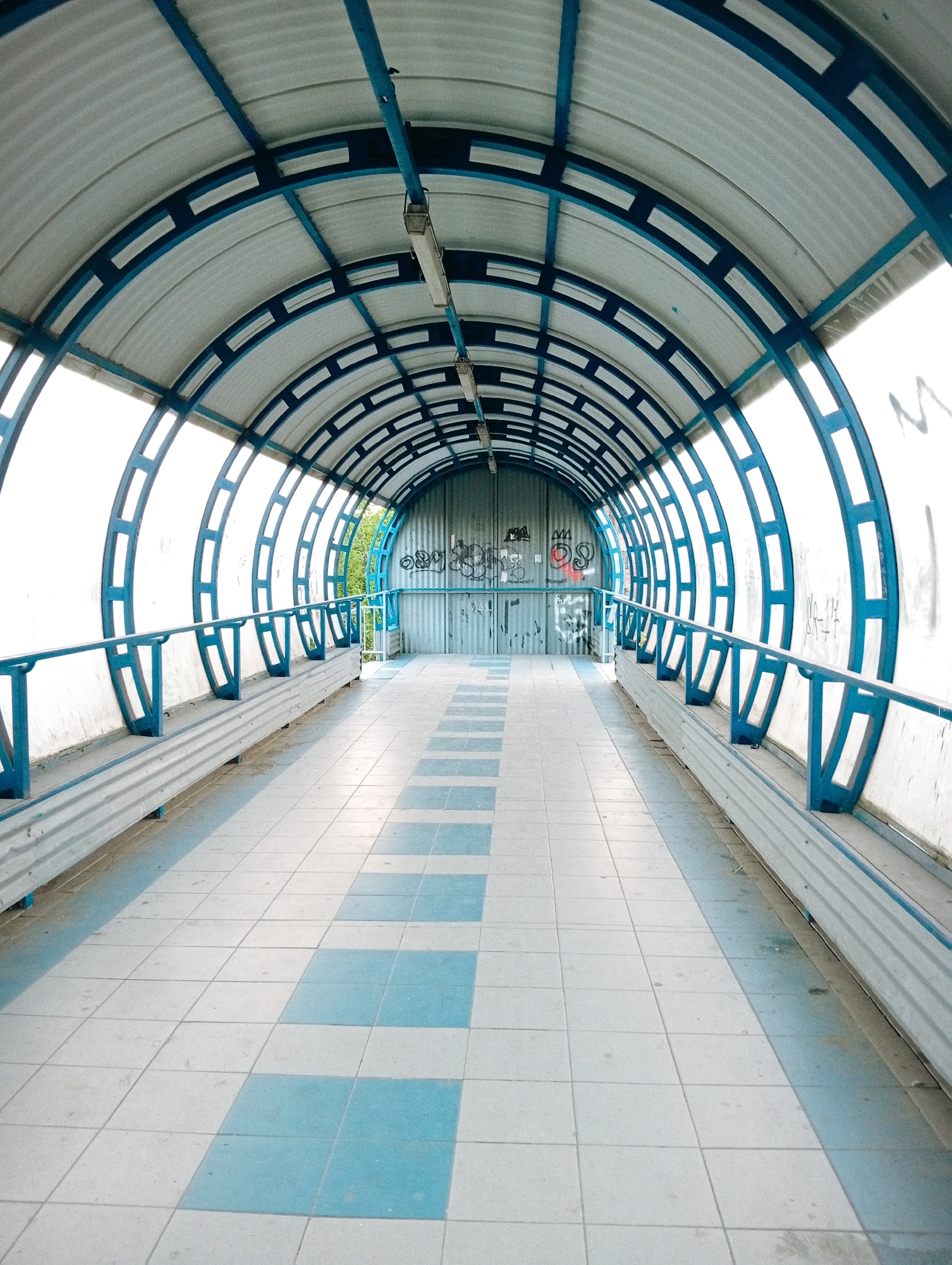 The image shows the interior of an elevated pedestrian walkway in Poltava, Ukraine. The walkway is covered by a blue-framed, arched metal structure, with translucent white panels forming the roof and walls. Fluorescent lights hang from the ceiling. Along each side, there are metal benches with blue railings. The floor is tiled in a checkered pattern of light blue and white. Graffiti is visible on the metal wall at the far end of the walkway. No people are visible in the scene.