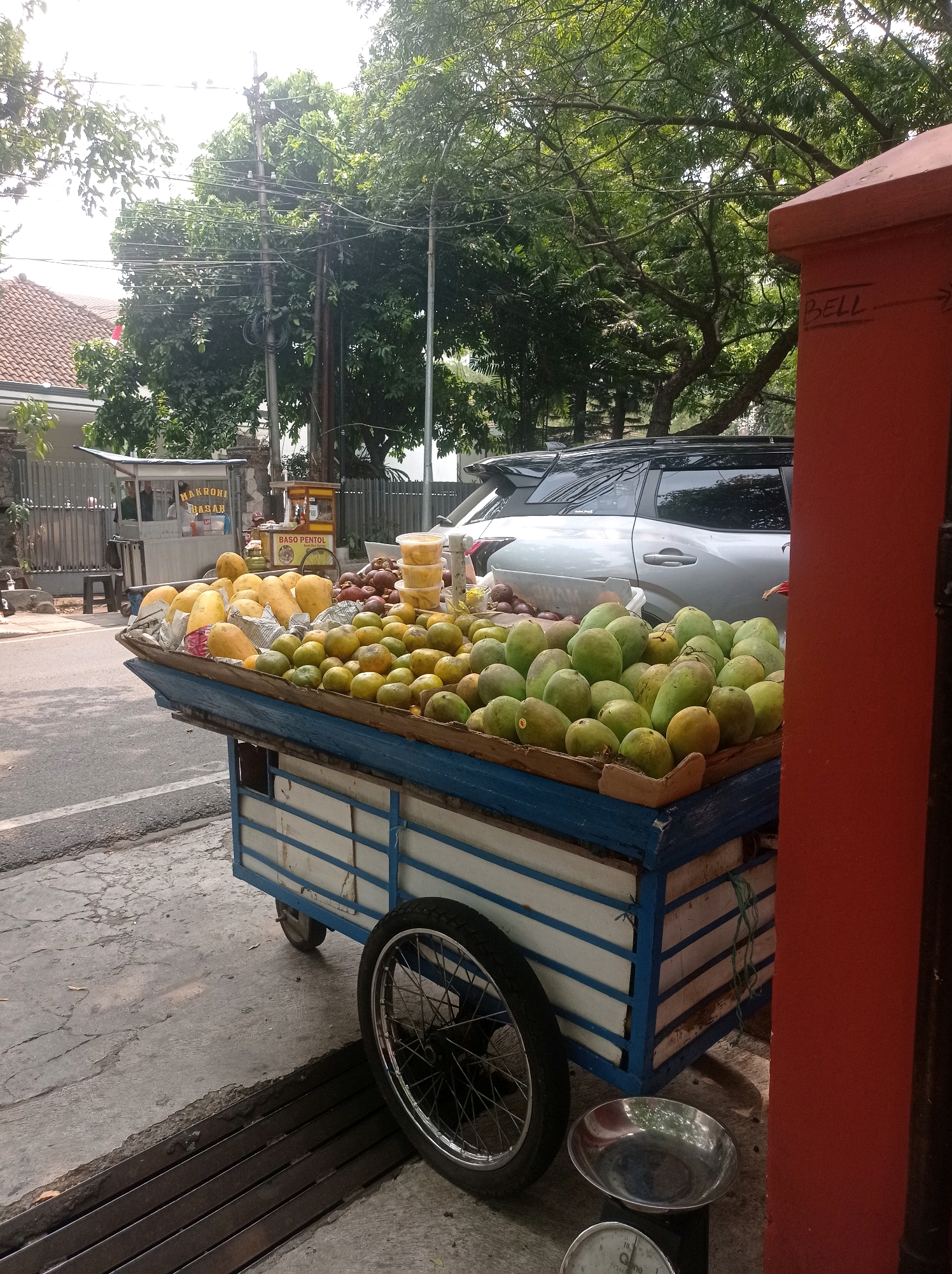 The image shows a fruit cart loaded with various fruits, including mangoes and oranges, on a street in Bandung City, Indonesia. The cart is blue and white with a wooden top. A silver car is parked behind the cart. Trees and a building are visible in the background. A red pillar is in the foreground. The scene appears to be a typical street market in Indonesia.