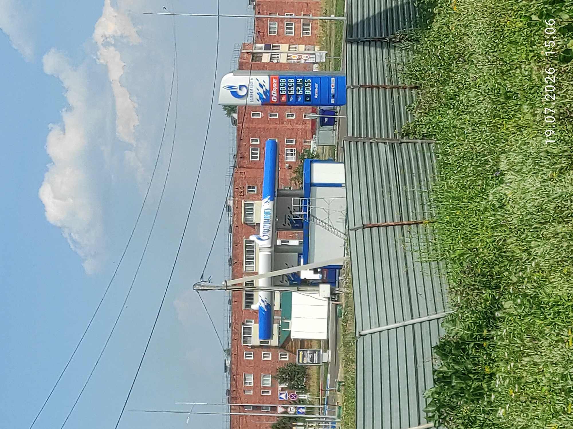 A gas station canopy is visible with a blue and white striped pattern. A tall sign displays prices for different types of gasoline. Behind the gas station are multi-story brick buildings with many windows. The sky is blue with some white clouds. In the foreground, a green metal fence runs horizontally across the image, with tall grass and weeds growing in front of it. The date and time "19.07.2026 15:06" are superimposed on the bottom right of the image.