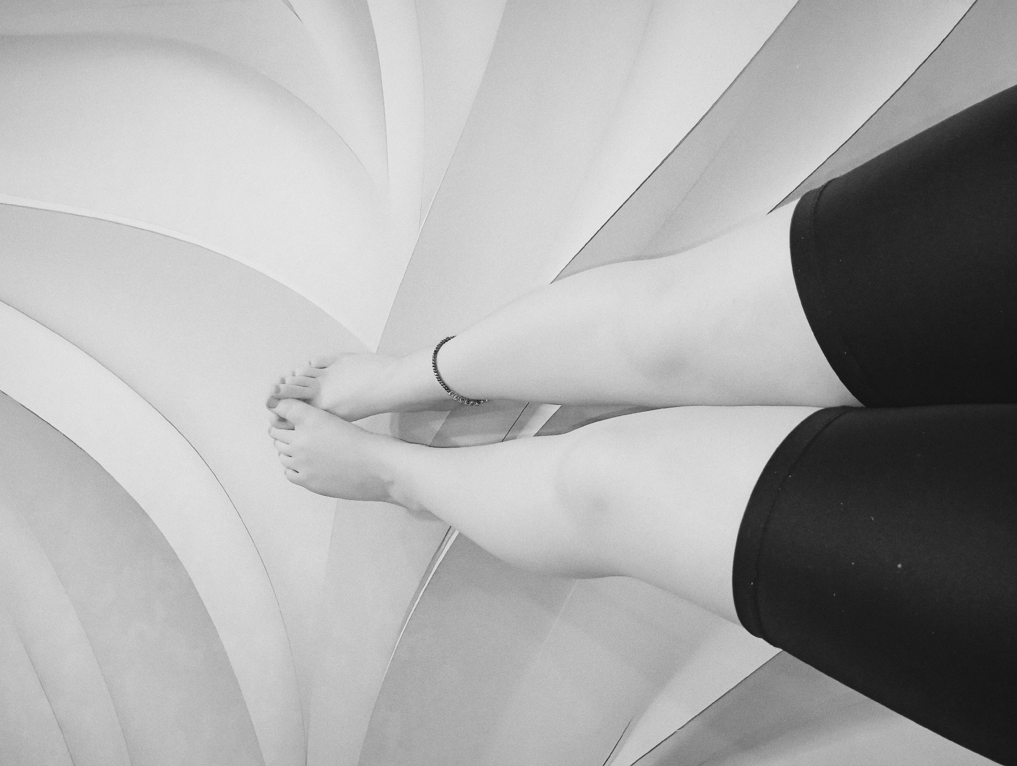 A black and white, high-angle, close-up shot depicts a person's legs and feet resting on a surface composed of layered, white and light gray segments. The person is wearing dark shorts that end mid-thigh. One leg is adorned with an ankle bracelet. The toes are visible on the feet. The surface appears to be a modern, architectural element composed of curvilinear shapes. The image was likely taken indoors. There is no way to ascertain the location is in Yurgamysh, Russia, based on the image alone.