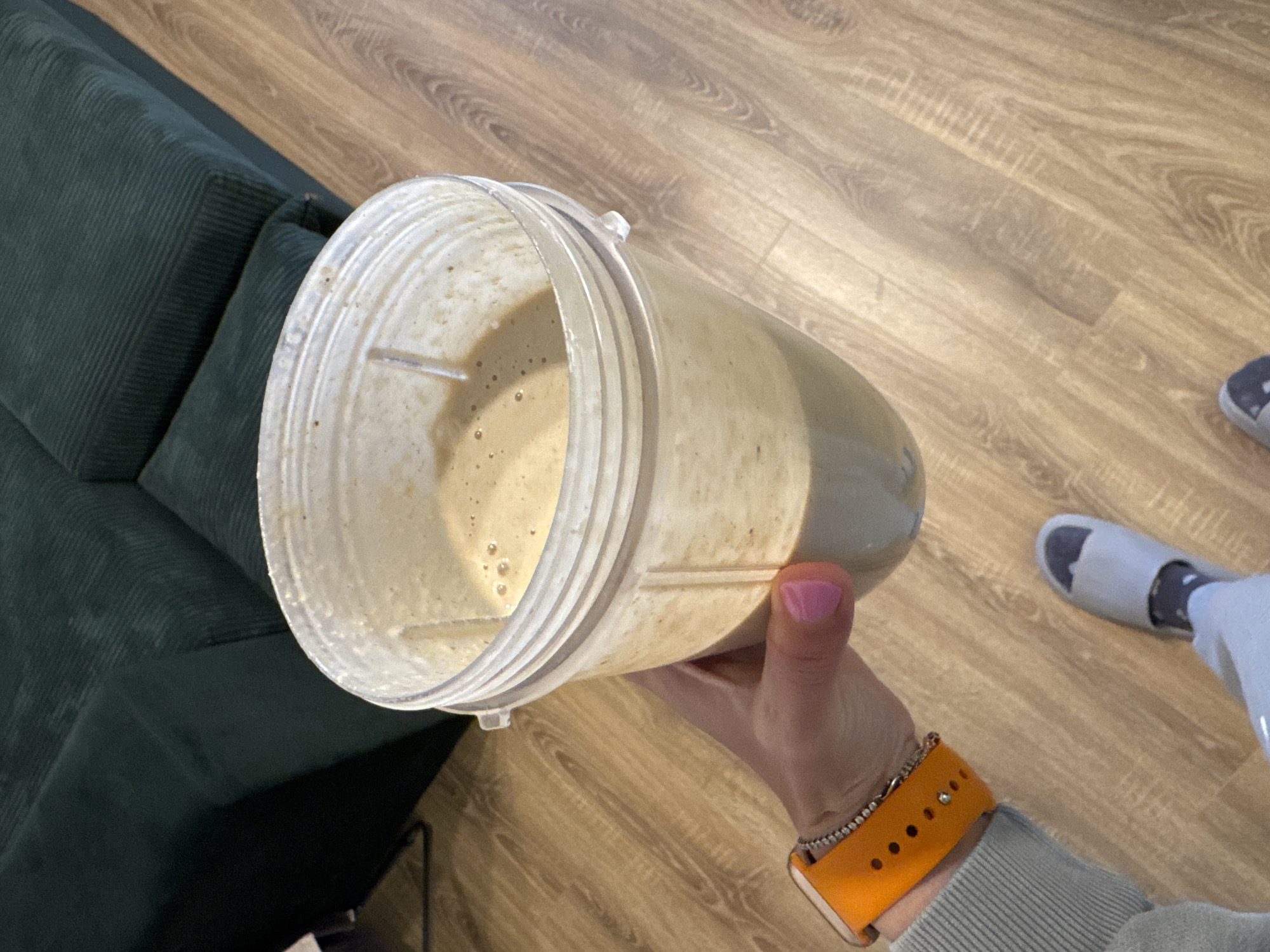 A first-person view shows a person holding a clear plastic blender jar with a beige-colored smoothie inside. The person is wearing a light grey long-sleeved shirt, an orange Apple Watch band with holes, and a silver bracelet on the left wrist. The fingernail on the thumb is painted pink. The blender is positioned above a light brown wooden floor. To the left, a dark green corduroy sofa is partially visible. In the background to the right, a white slide with a dark grey sock inside is visible. The location is indoors. There is no evidence to suggest this image was taken in Sofia, Bulgaria.