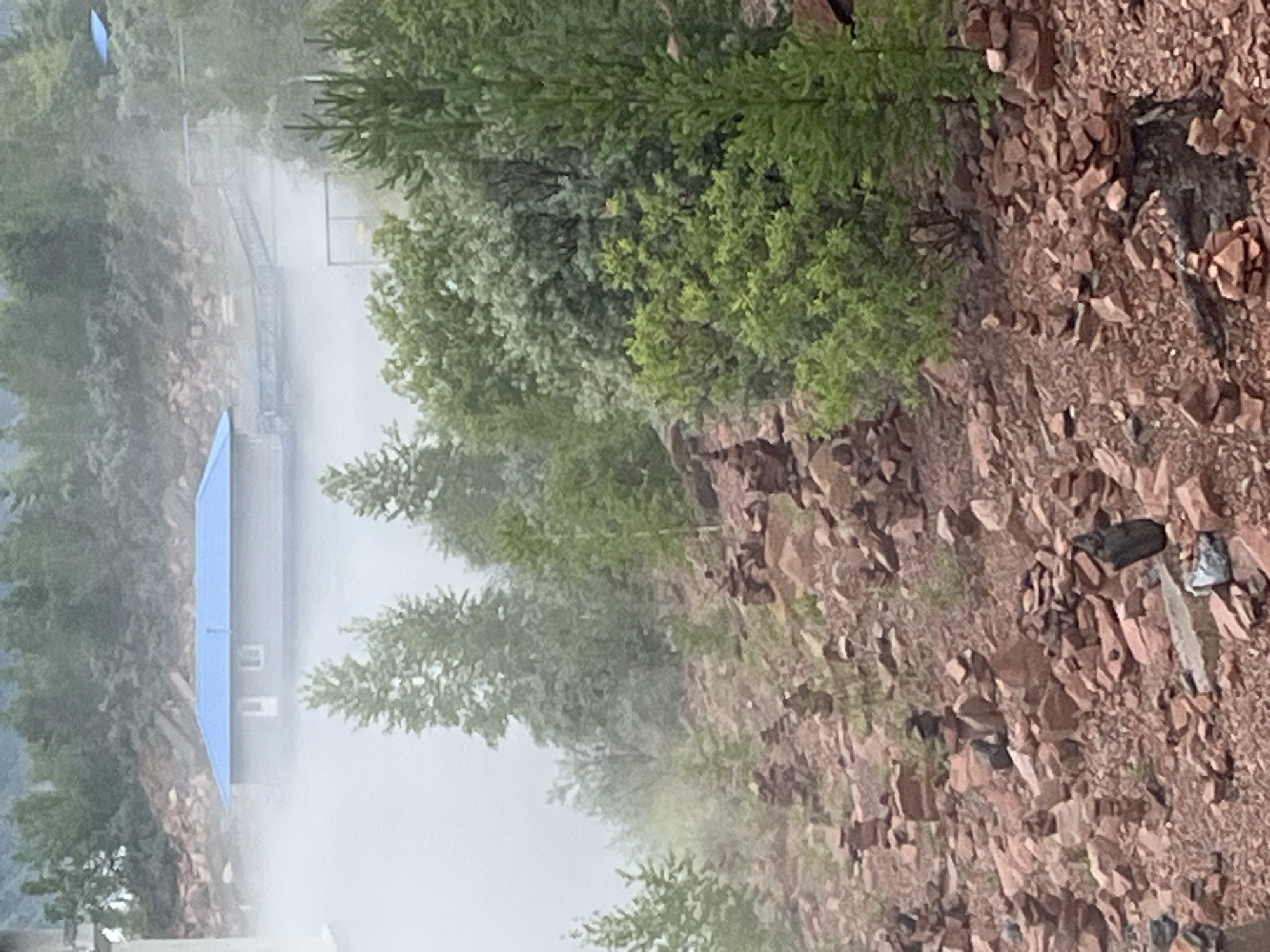 The image depicts a misty, mountainous landscape near Divnogorsk, Russia, characterized by an abundance of stacked rock cairns scattered across a reddish-brown, rocky terrain. In the background, a structure with a blue roof is visible, seemingly floating on water and partially obscured by fog. Several trees, some appearing evergreen, are interspersed among the rocks and in the background. The overall atmosphere is damp and hazy. No people or animals are visible in the image. The stacked rocks suggest a human presence and possibly a trail or spiritual site. The presence of the blue-roofed structure and a bridge-like element suggests a man-made area, perhaps a recreational or industrial site, within the natural mountainous environment.