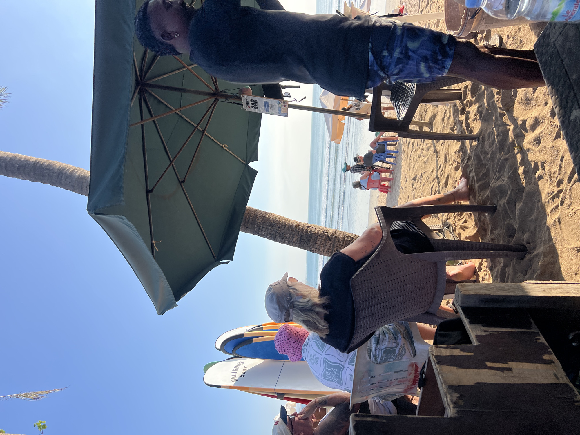 In Kuta, Indonesia, a beach scene unfolds under a clear blue sky, with gentle waves lapping the shore. In the foreground, several individuals are seated on chairs on a sandy beach, some under the shade of a large green umbrella. A person with dark skin and a mohawk hairstyle, wearing blue patterned shorts and a black t-shirt, stands on the right, facing away from the camera. Beach umbrellas and surfboards, one with "BALIGUNS SURF" written on it, are visible. In the background, a few more people are seated on colorful chairs near the water's edge, and a few more beach umbrellas are set up. The ocean stretches to the horizon, with a calm sea and a few small waves. A palm tree trunk is partially visible behind the umbrella. A sign for "CHERRY BAR" hangs from the umbrella.