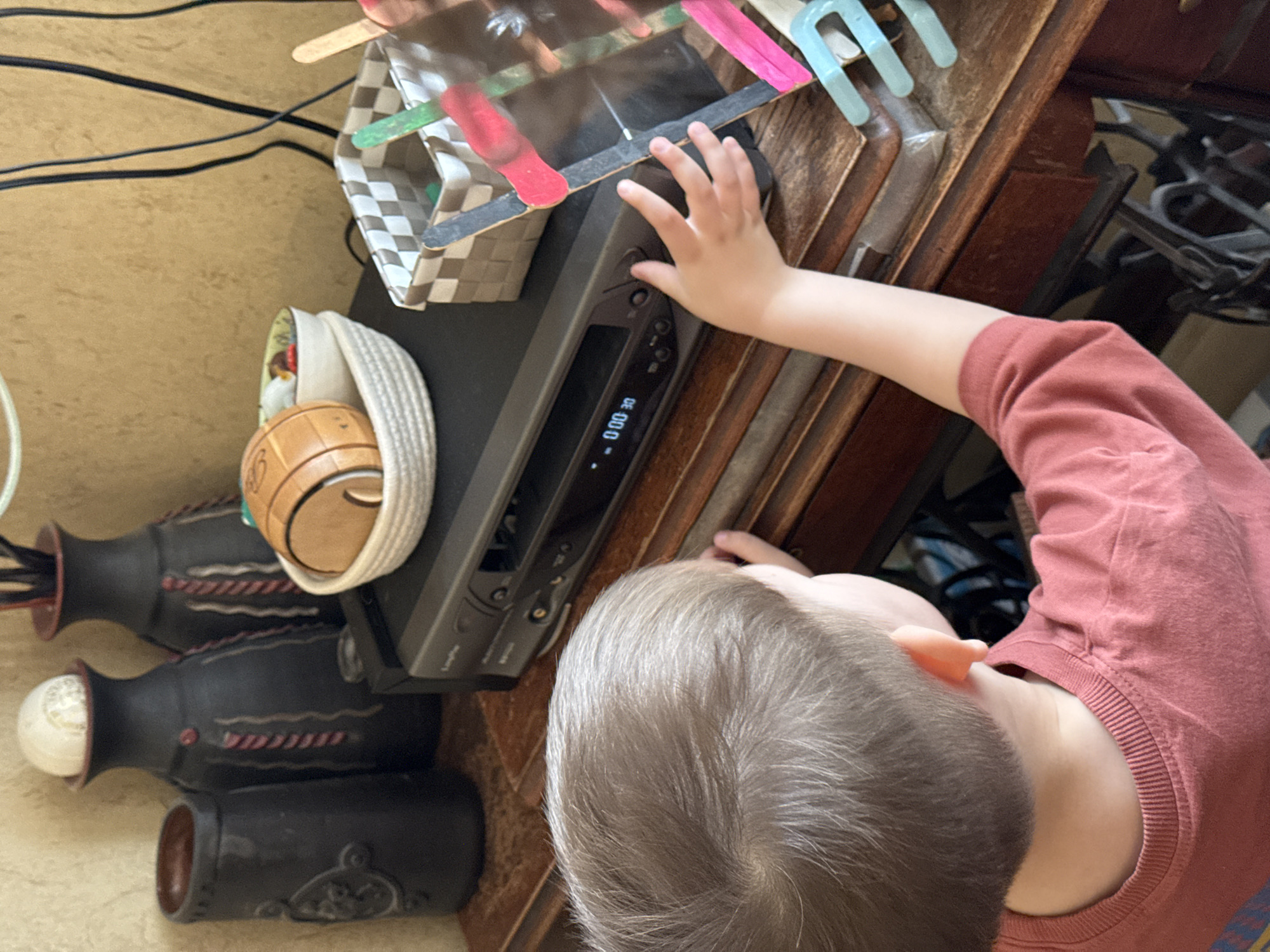 A high-angle shot depicts a young child, with light-colored hair and wearing a maroon shirt, reaching towards a black electronic device with a digital display showing "00:00.0". The device sits on a wooden surface, possibly a cabinet or shelf. To the left of the device are decorative objects, including three black vases with white and red accents, a small wicker basket containing what appears to be a small ball, and a homemade container constructed from popsicle sticks. Electrical cords are visible in the background. There is no information in the image to suggest the location is in Moscow, Russia.