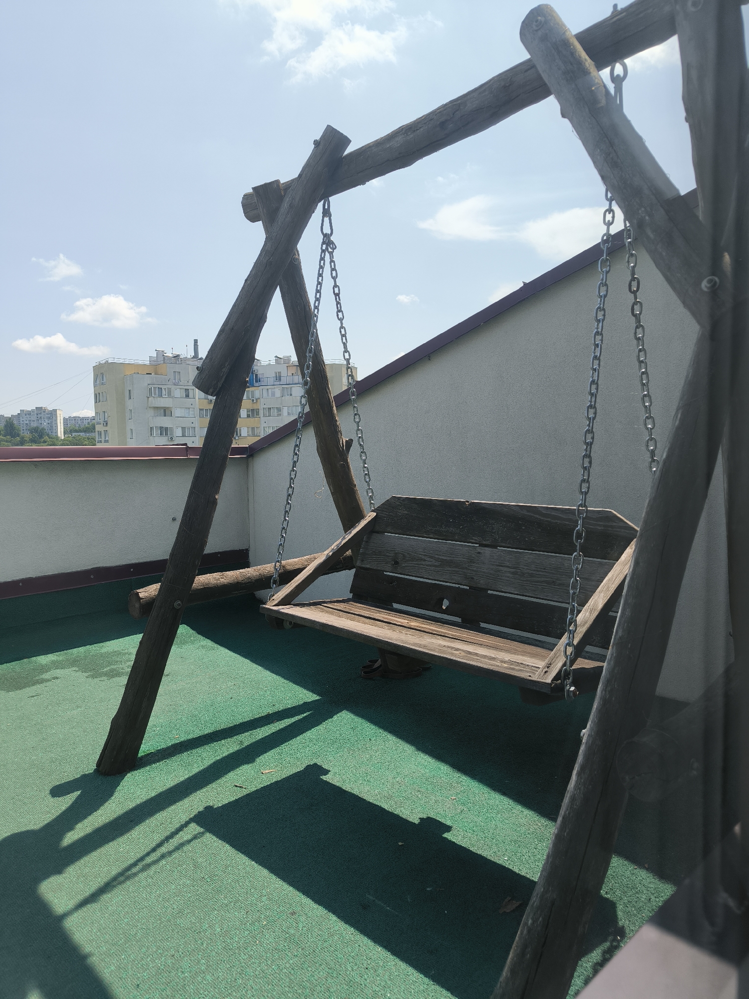 The image shows a wooden swing set on a green surface, possibly a rooftop terrace, in Chișinău, Moldova. The swing set has a wooden bench suspended by chains from a wooden frame. In the background, there are apartment buildings and a clear blue sky with scattered clouds. The sun casts long shadows on the green surface, indicating it is daytime. The overall scene is tranquil and suggests a residential setting.