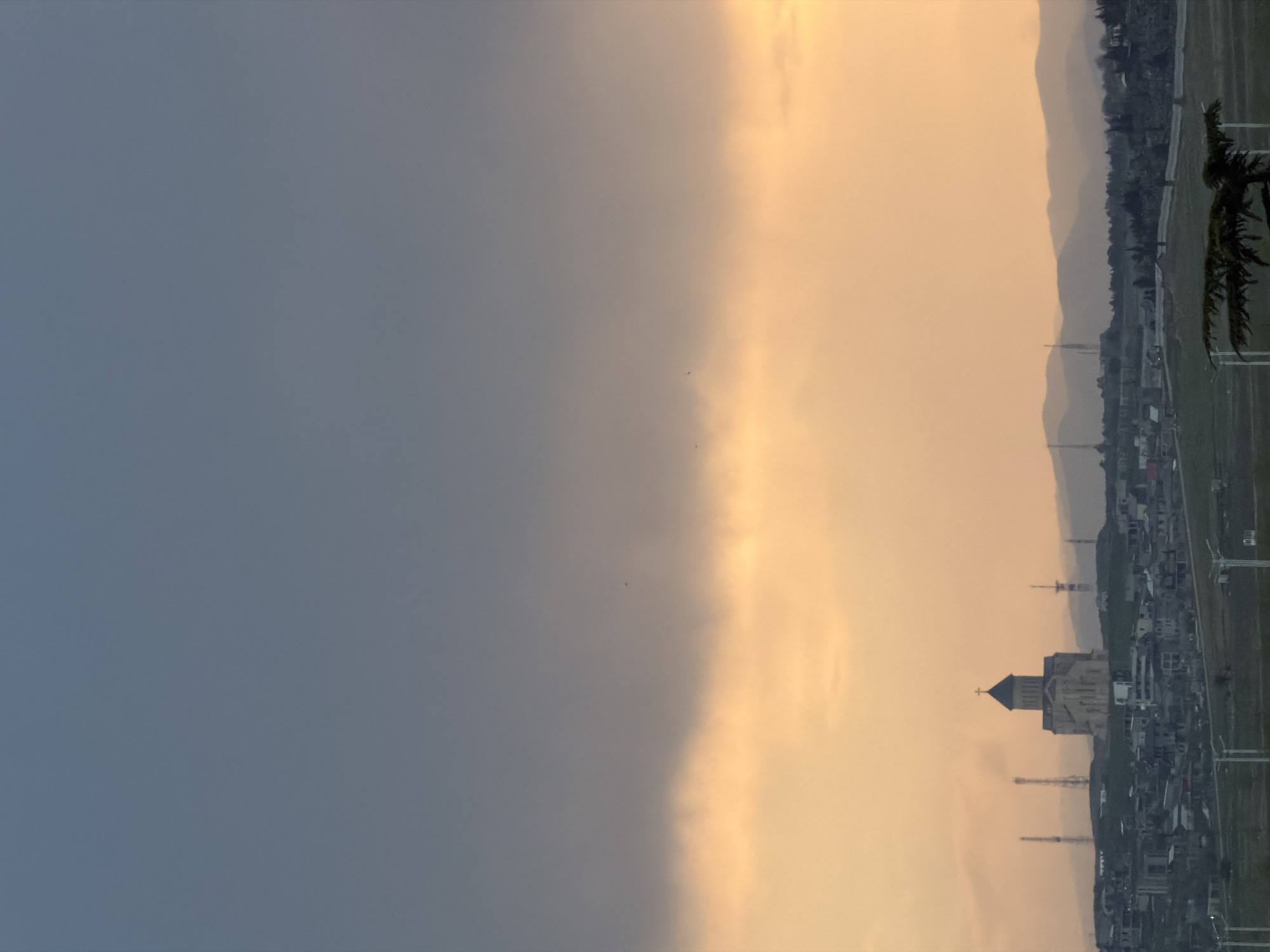 The image presents a wide landscape view of a city or town backed by mountains, under a dramatically divided sky. The sky is split into two distinct zones: a dark, deep blue-grey occupying the left side of the frame, and a vibrant golden-orange on the right, suggesting either sunrise or sunset. A clear, somewhat jagged line marks the transition between these two atmospheric conditions, indicative of a weather front or specific cloud illumination.
Below the illuminated sky on the right, a cityscape unfolds. A prominent structure resembling a stone church or cathedral, featuring a dark, tiered roof and a cross at its highest point, stands out in the midground. Surrounding it are various other buildings, possibly residential or commercial, contributing to the urban sprawl. Several tall, slender transmission towers or communication masts are scattered across the city and visible against the horizon.
In the background, a series of layered mountains or hills stretch across the scene, appearing hazy and soft against the sky. On the far right, a dark green coniferous tree or shrub partially enters the frame. Tiny specks, likely birds, are faintly visible in the sky near the dividing line of the clouds. The dramatic lighting and contrasting skies create a striking and atmospheric view of what, according to context, is Tbilisi, Georgia. No text is visible in the image.