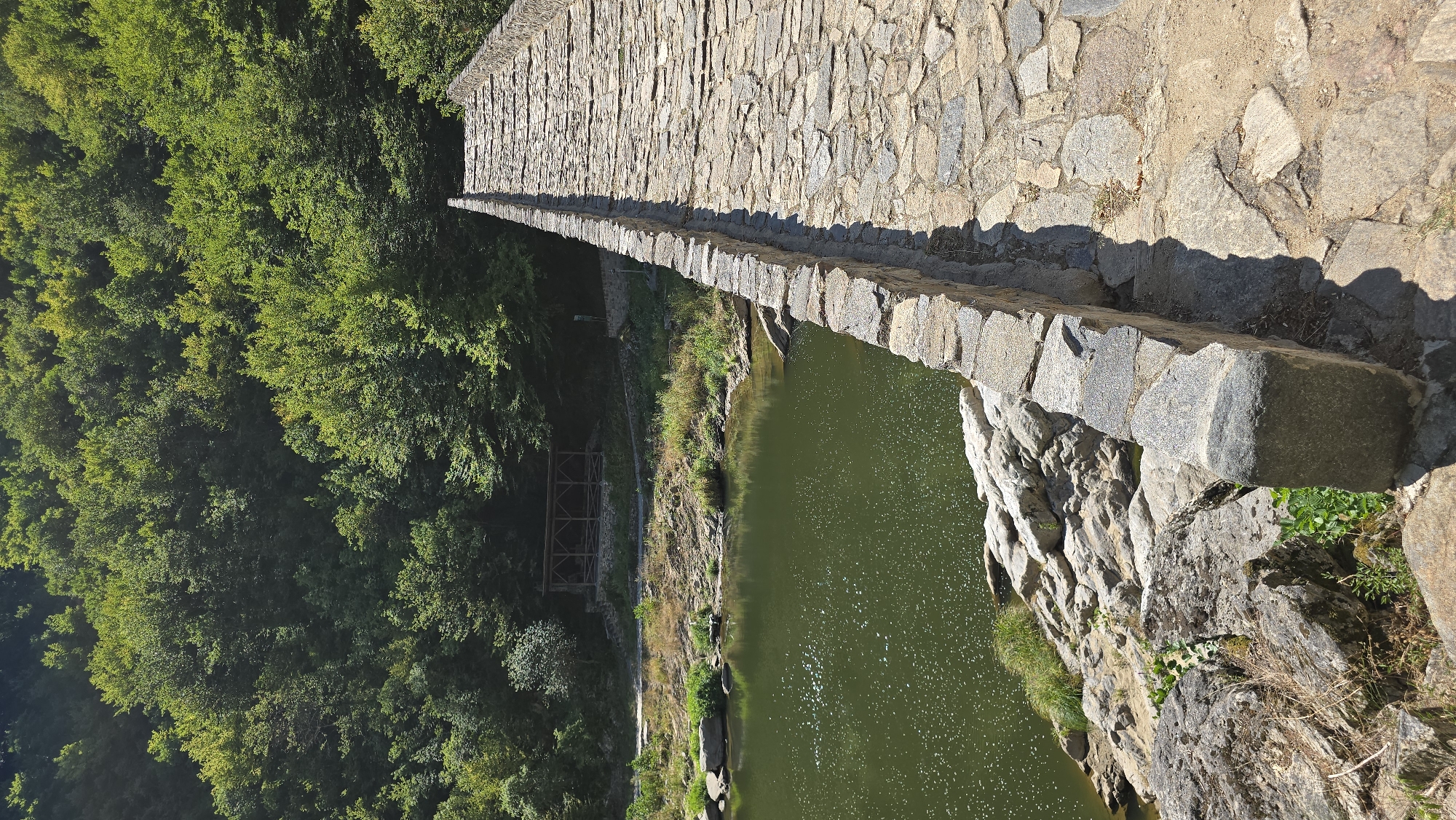 The image depicts a stone bridge arching over a green body of water, likely a river, in Diadovtsi, Bulgaria. The bridge is constructed from rough-hewn stones, with a worn pathway forming the surface. The water below is calm, reflecting the sunlight and the surrounding lush green foliage. On the left bank, rocky outcrops and vegetation lead to a wooded hillside densely packed with trees. A wooden structure, possibly a gazebo or rest area, is visible nestled among the trees on the bank. The perspective is from the surface of the bridge, looking forward along its length towards the distant treeline. The scene is bathed in natural daylight, casting shadows across the stone pathway. There are no people visible in the image.