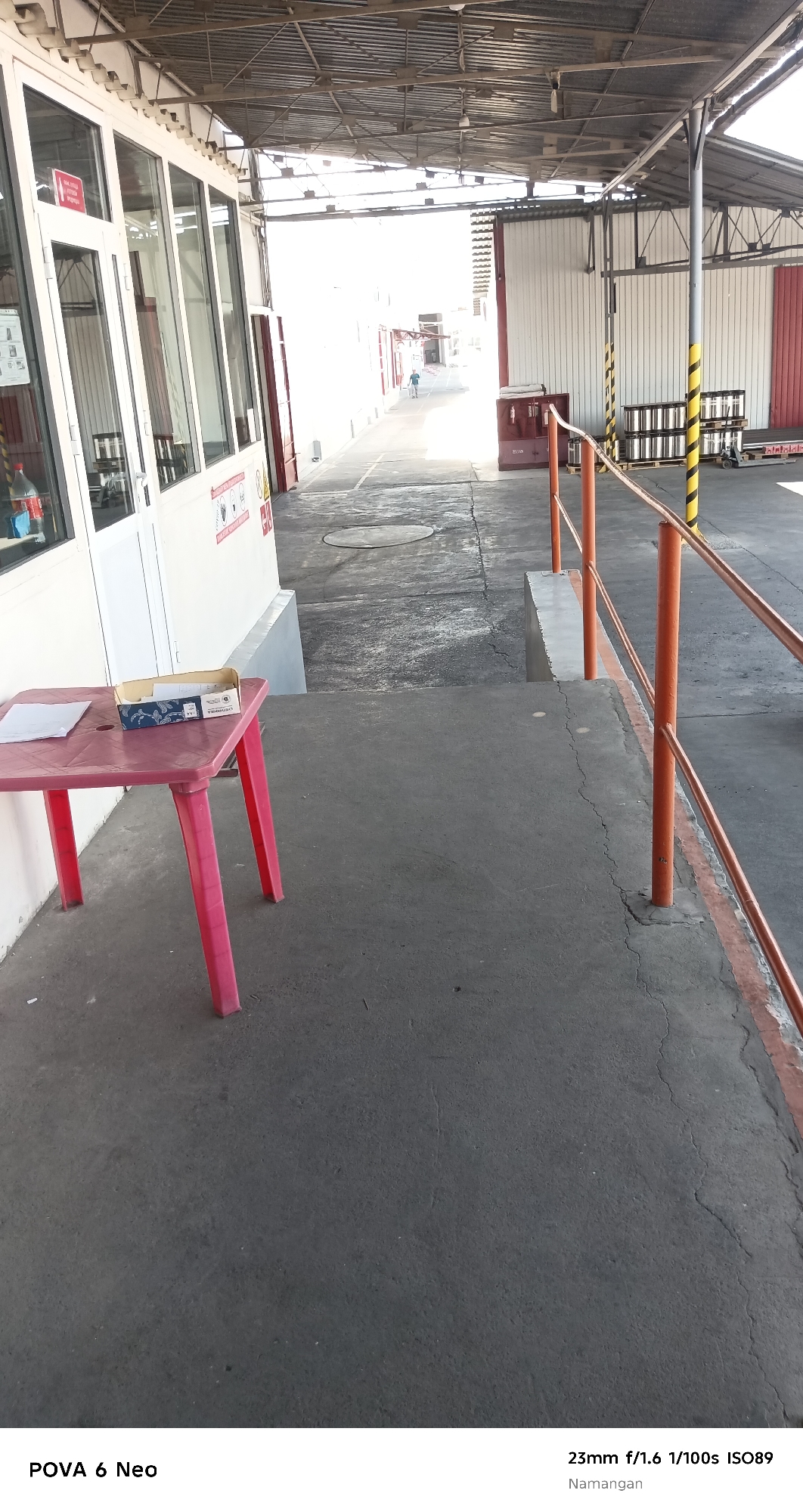 The image depicts an outdoor industrial or storage area, likely within the city of Namangan, Uzbekistan, as indicated by the text overlay. In the foreground, a small, bright red plastic table stands on a concrete surface. On the table, there is a white piece of paper and an open, blue and white cardboard box. A red bottle with a white label is visible in the background on the left side.
The environment appears to be a walkway or service area adjacent to a building with large glass windows and white siding. A metal railing with orange posts and handrails runs along the right side of the walkway, separating it from a slightly elevated concrete platform. Beyond this, industrial structures and possibly loading docks are visible, characterized by metal roofing, corrugated metal siding in white and red, and stacked pallets. A person, wearing a blue shirt and light-colored shorts, is walking in the distance down the pathway, seemingly in a less illuminated area. The overall lighting suggests it is daytime, with bright sunlight illuminating the far end of the area, creating strong contrast. The ground is predominantly concrete, showing signs of wear and some cracks. The text overlay at the bottom of the image provides camera details: "POVA 6 Neo" and "23mm f/1.6 1/100s ISO89", along with the location "Namangan".
