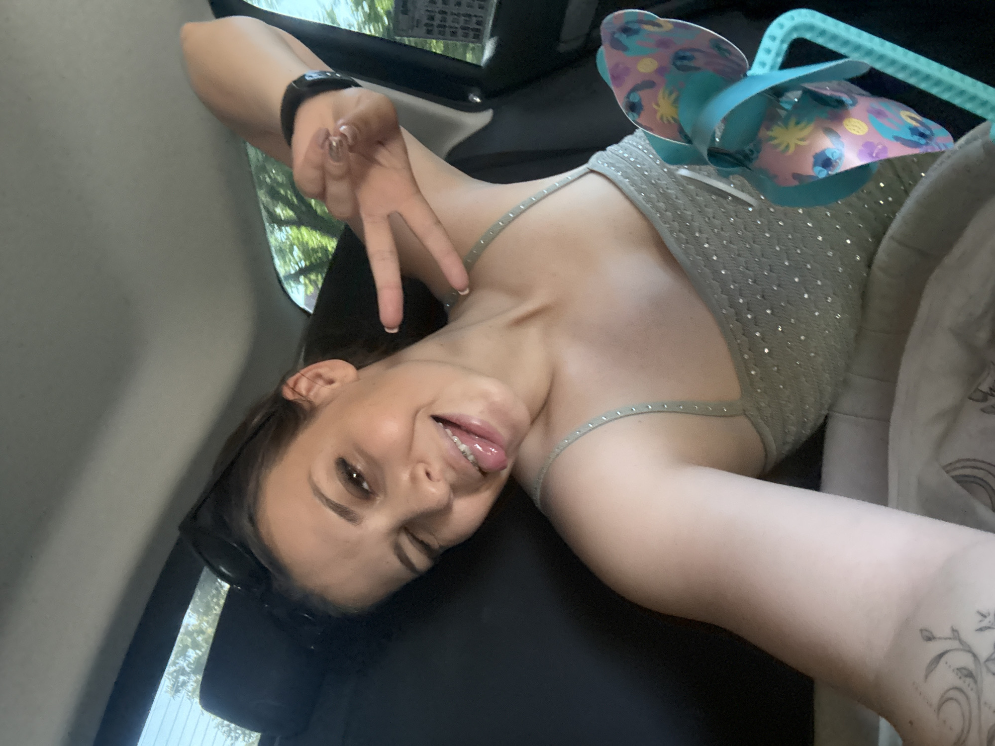 A young woman is lying down inside a vehicle, possibly a car, with her body angled to the right. She is wearing a sparkling light green tank top with thin straps adorned with small rhinestones. Her left arm is raised and bent at the elbow, with her hand extended and making a peace sign with her index and middle fingers. She is wearing a black fitness tracker on her left wrist and has long, light brown nails.
Her head is tilted, and she is looking towards the camera with one eye winking and her tongue playfully sticking out. She has dark brown hair that is partially visible and dark eyebrows. Sunglasses are resting on her nose.
In the background, a light blue toy with colorful patterns, resembling a pinwheel, is attached to a turquoise plastic handle. The interior of the vehicle appears to be dark, likely a seat or a car interior lining. Through the windows, greenery and trees are visible, suggesting an outdoor setting on a sunny day. The overall mood conveyed is playful and energetic.