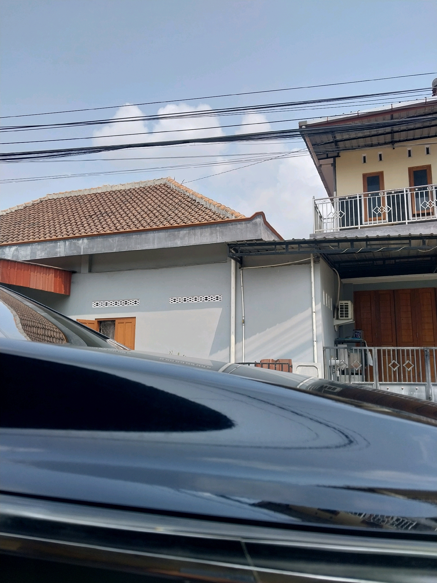 This is a medium-angle, outdoor shot in the city of Purwomartani, Indonesia, under a clear blue sky with some wispy clouds. The foreground is dominated by the highly reflective, dark, curved surface of a car, which mirrors the sky and the surrounding environment. In the midground, a grey-walled building with a tiled roof is visible on the left, featuring brown-framed windows and decorative vent openings. To the right of this building is a two-story structure with a light yellow upper facade, featuring windows and a metal railing on the balcony. The lower level of this building has large, dark wooden doors and a metal fence. Overhead, multiple black electrical wires crisscross the scene, supported by poles not visible in the frame. An air conditioning unit is mounted on the wall of the yellow building. The overall impression is a residential street scene in Indonesia.