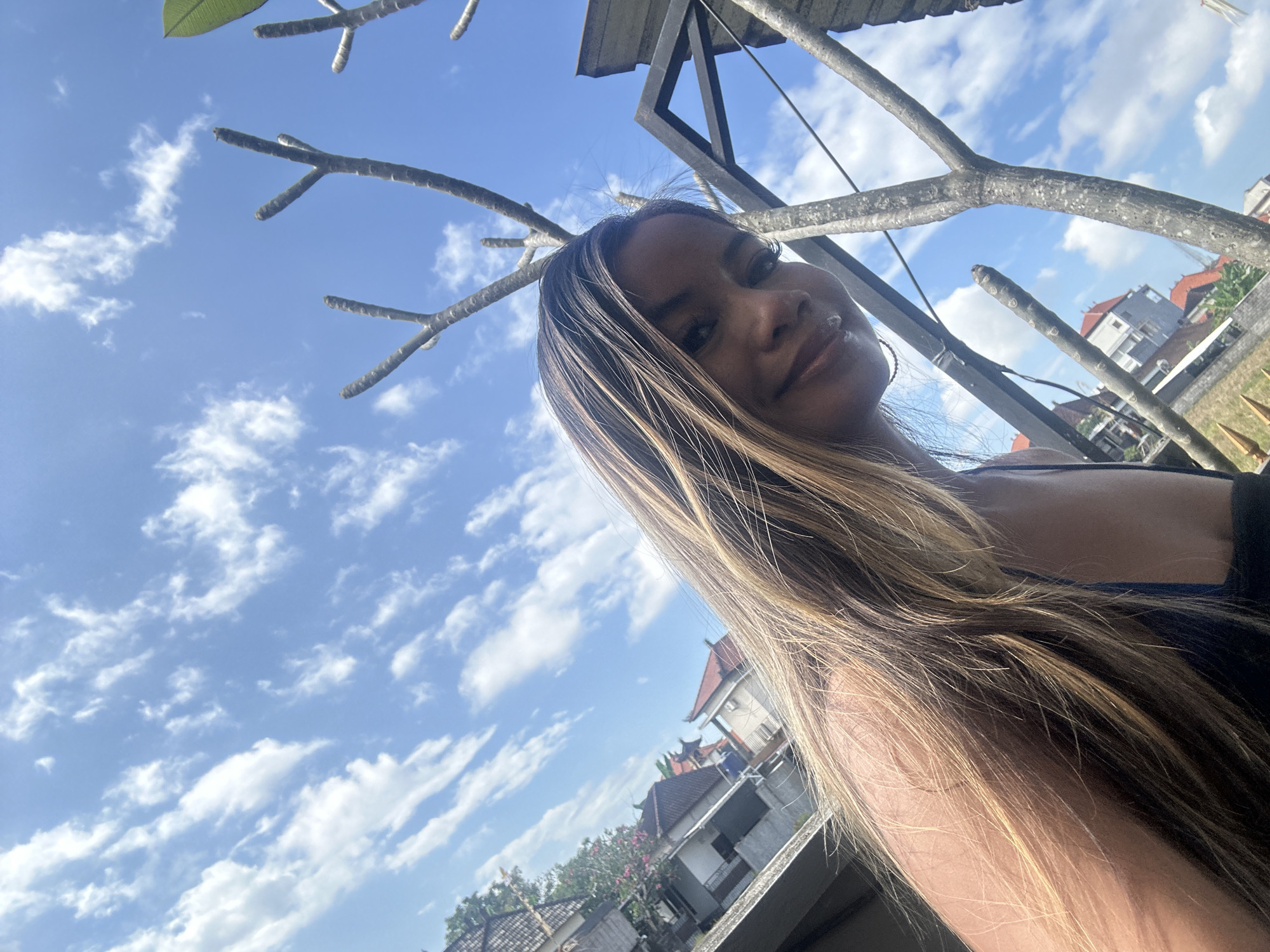 This is a selfie taken outdoors during the day, likely in Sukawati, Indonesia. The image is angled upwards, with a bright blue sky filled with scattered white clouds dominating the background. In the foreground, a person is visible, looking towards the camera with a slight smile. Their long, blonde-streaked hair falls forward, partially obscuring their face. Bare branches of a tree extend across the frame, and a portion of a dark structure, possibly a roof or awning, is visible at the top. In the distance, a cluster of buildings with red tiled roofs can be seen, typical of Indonesian architecture. The sunlight suggests it is daytime, and the clear sky with clouds indicates pleasant weather. The overall mood appears relaxed and cheerful. No visible text is present in the image.