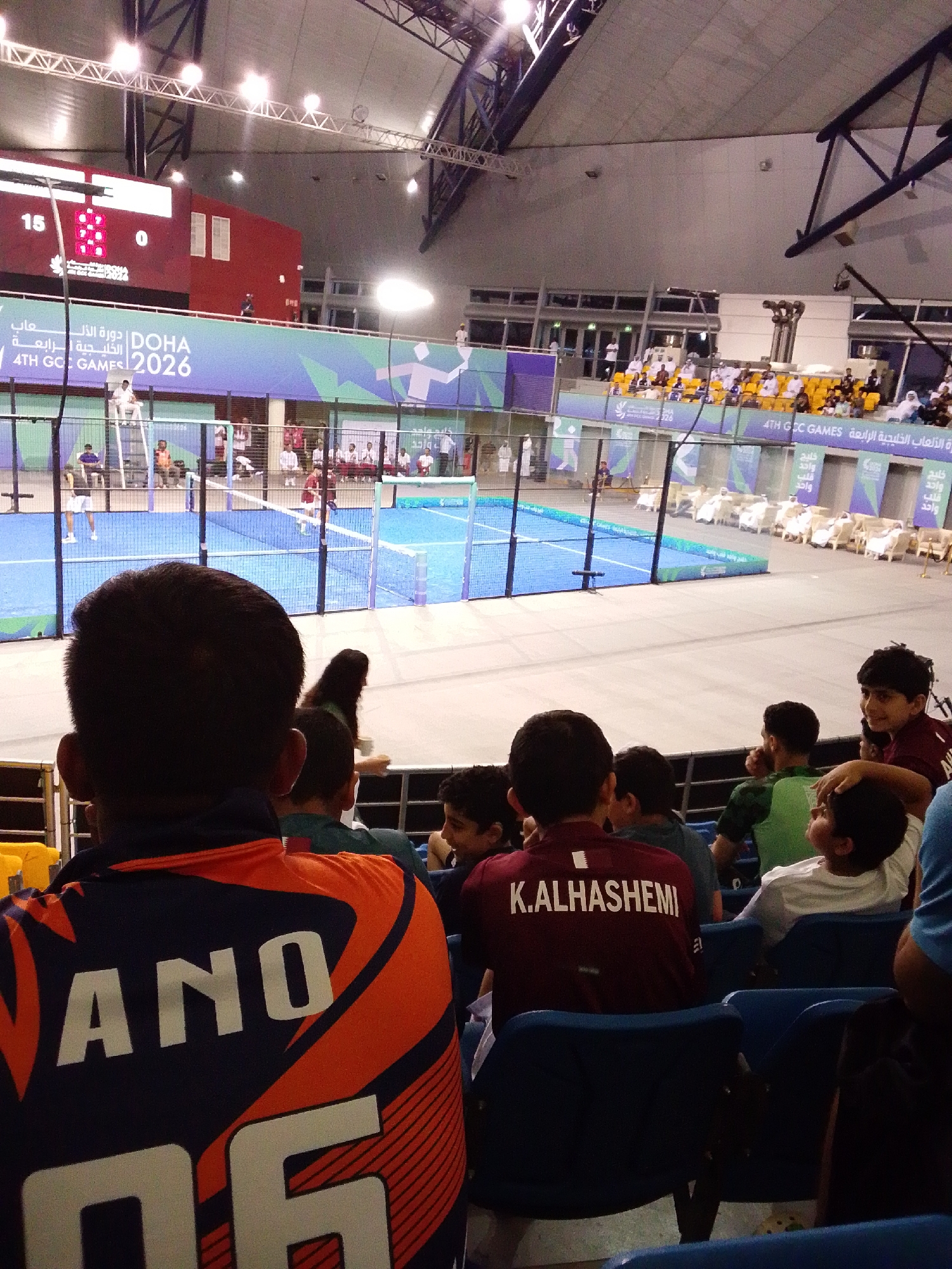 Here is a detailed description of the image:
**Scene:** The image captures a sporting event, specifically a padel tennis match, held indoors in Al Rayyan, Qatar. The event is part of the 4th GCC Games 2026, as indicated by the banner and signage visible in the background.
**People:** The image shows spectators watching the padel tennis match. In the foreground, several children are seated in blue stadium chairs. The back of one child's jersey reads "ANO 06," while another child's jersey displays "K.ALHASHEMI."
**Subject:** The main subject of the image is the padel tennis match. Two players are visible on the blue padel tennis court, surrounded by a wired fence and a net in the middle.
**Objects:**
* A large scoreboard displays the score.
* Banners promoting the "DOHA 4TH GCC GAMES 2026."
* Stadium seating in various colors, including yellow and blue.
* A net between the two sides of the court.
**Actions:** The primary action is the padel tennis match in progress. Spectators, including the children in the foreground, are watching the game.
**Interactions:** The spectators are engaged in watching the padel tennis match.
**Environment:** The event is taking place in an indoor stadium with a high ceiling and metal framework. The lighting is bright, illuminating the padel tennis court and the surrounding spectator areas.
In summary, the image shows spectators, including children, watching a padel tennis match during the 4th GCC Games 2026 in Al Rayyan, Qatar.