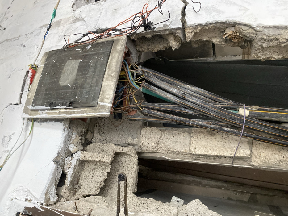 The image displays a severely damaged interior wall within a building in Khan Yunis, Palestinian Territory. A partially dislodged electrical distribution panel, featuring a dark, rectangular closed door, is mounted on the wall. Below and behind the panel, a dense cluster of multi-colored electrical wires (including yellow, blue, orange, brown, and green) is exposed. A bundle of approximately ten black plastic conduits or protective sleeves, some containing wires, hangs vertically downwards from the opening beneath the panel. Loose orange and black wires are visible extending from the right side of the panel. The surrounding wall exhibits extensive damage, with large sections of white plaster and concrete missing, revealing fractured grey cinder blocks and raw concrete debris. Two bent metal rebar rods protrude from the damaged section on the left. Numerous cracks are visible across the remaining plaster surfaces. No individuals are present in the scene.