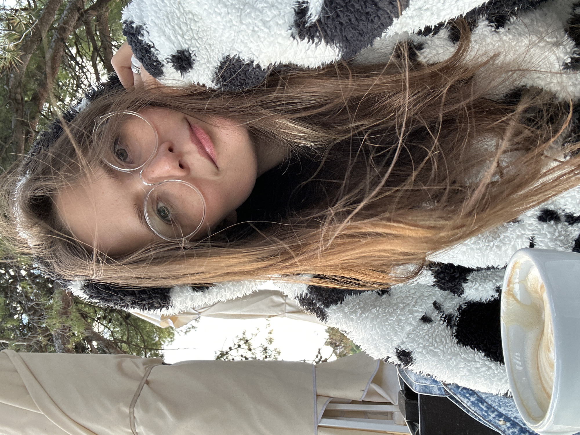 A young woman with long brown hair and round glasses is wearing a black and white cow-patterned hooded jacket. She appears to be taking a selfie. A white mug containing a latte is visible to the lower right of the image. Trees are visible in the background. The image was taken outdoors, possibly in Šušanj, Montenegro.