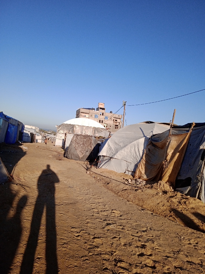 The image displays a ground-level view of a makeshift camp or informal settlement in Khan Yunis, Palestinian Territory, under a clear, cloudless blue sky. A wide, packed dirt path, uneven and marked with numerous footprints, occupies the foreground and extends into the midground. Two distinct, elongated human shadows are cast onto the path from the left side. On both sides of the path, rows of temporary shelters are visible, constructed from various materials including light grey and blue tarpaulins, brown fabrics, and exposed wooden support frames; some shelters have earth mounded around their bases. In the background, a multi-story concrete building with visible damage or boarded windows and a rooftop water tank is prominent. A smaller white-domed structure is positioned closer to the background building. A wooden utility pole supporting a light fixture and overhead power lines stands to the right, extending towards the distant structures.