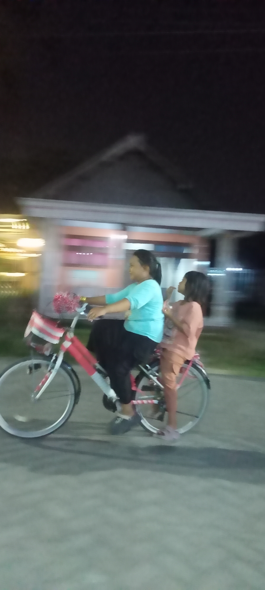 An over-the-shoulder, low-angle, motion-blurred photograph taken at night shows two individuals on a bicycle on a paved road in Gempolklutuk, Indonesia. The primary subject is a woman wearing a turquoise shirt and black pants, riding the bicycle and facing right. She is holding onto the handlebars, which are decorated with red and white streamers. A younger girl in a peach-colored shirt and shorts is seated behind the woman on the rear rack of the bicycle, facing left and appearing to talk to the woman. The bicycle is red and white. In the background, a building with a pitched roof is visible, with a row of lit windows. The ground is a light gray paved surface with shadows. The image captures motion with streaks of light and blurred figures.