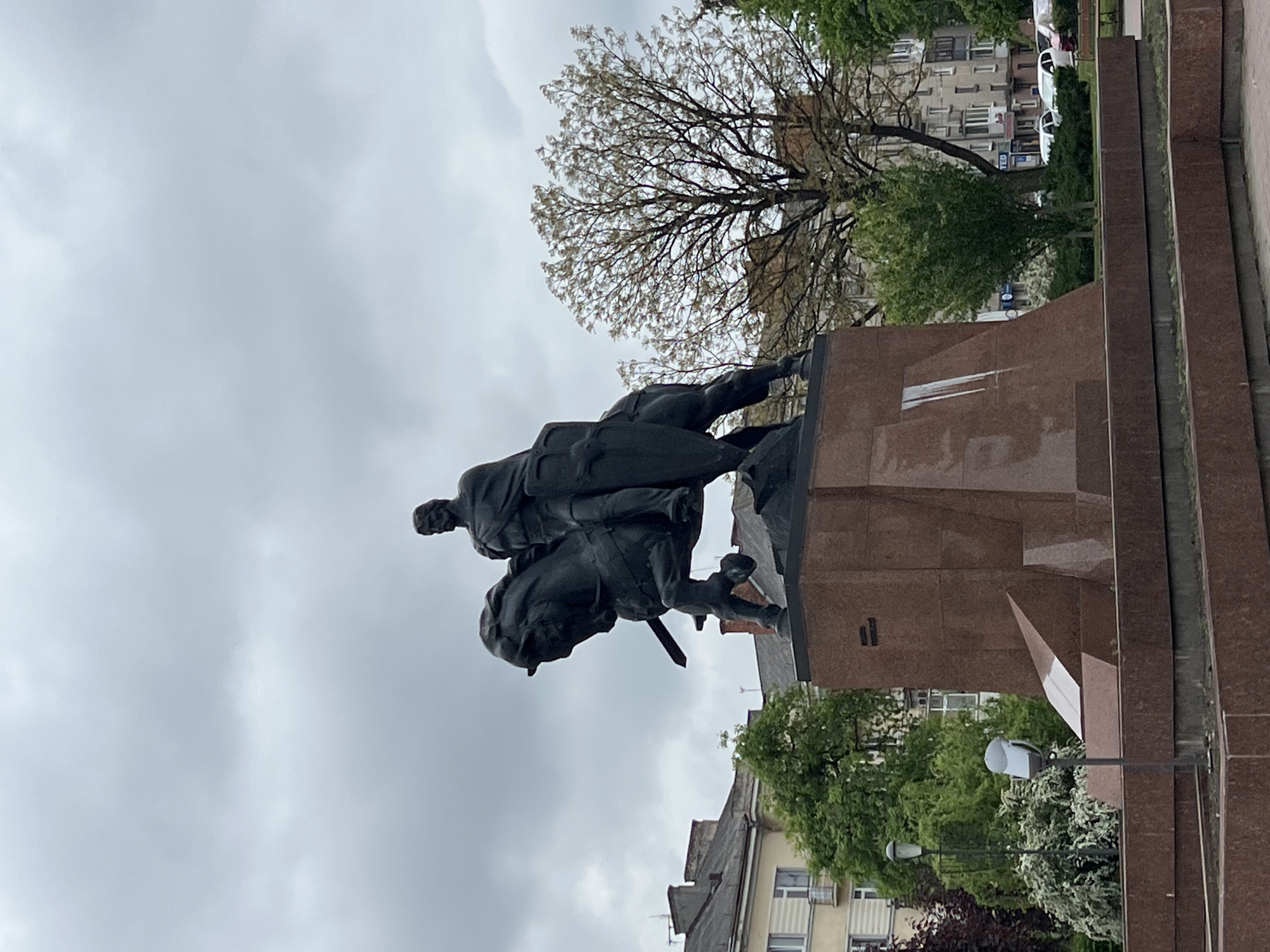 The image shows a statue of a man on horseback, mounted on a tall, brown stone pedestal. The statue is dark in color, contrasting with the overcast sky. The statue is positioned in front of a background of buildings and trees. In the background, there are several buildings, including a building with a cream-colored facade and a building with a gray roof. The trees are green and leafy. The statue is the Taras Shevchenko Monument in Ternopil, Ukraine.