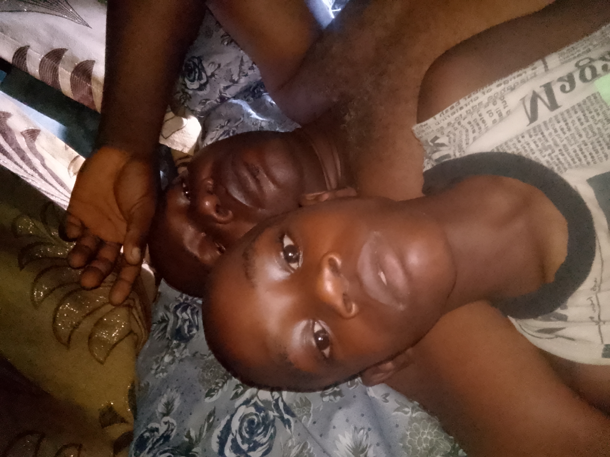 Two dark-skinned individuals are lying on a bed. The person in the foreground has their head turned toward the camera, while the person in the background is partially obscured. The bed is covered with patterned sheets. The location is Unknown, Unknown.