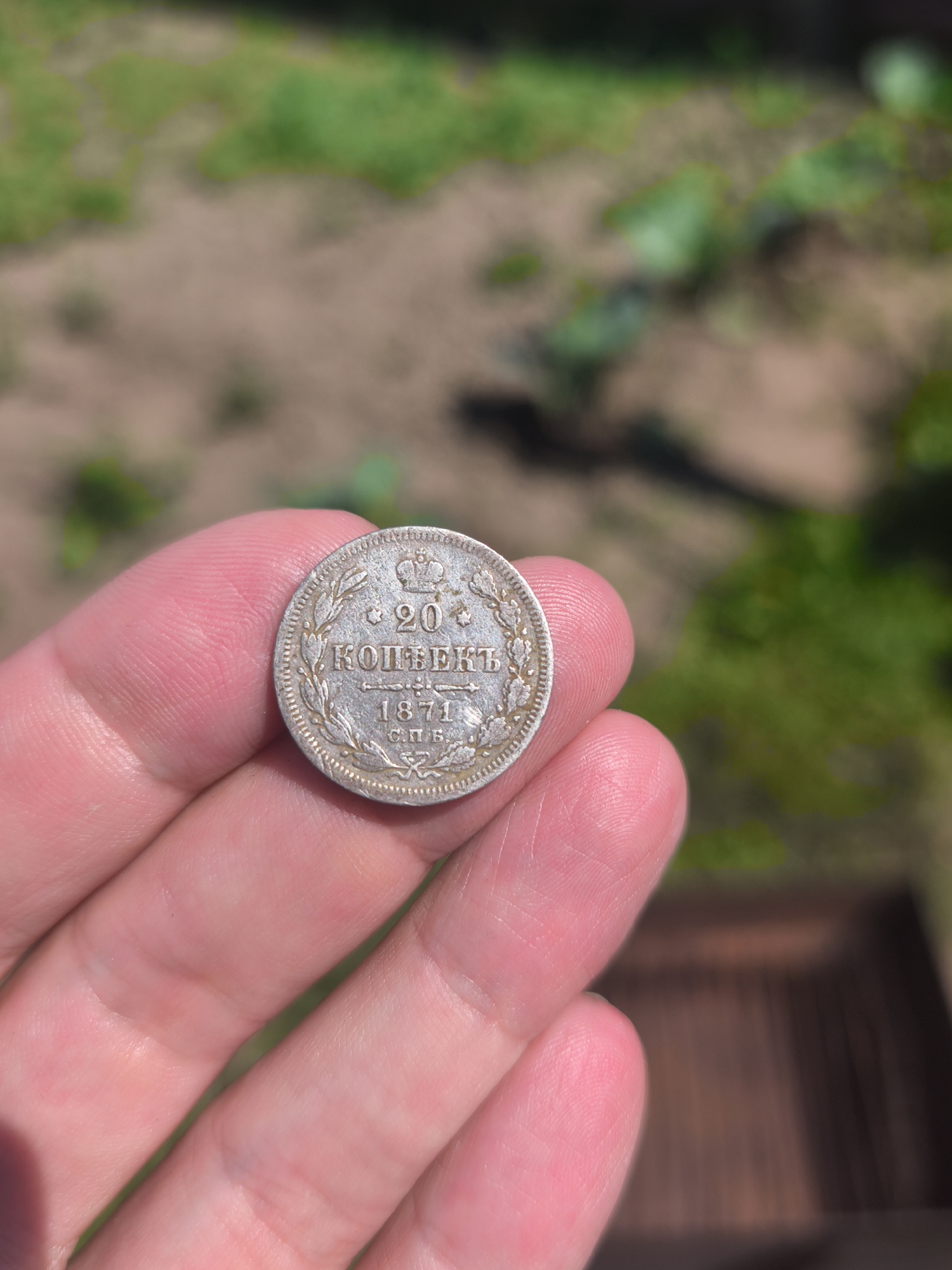 A hand holds a silver coin with Cyrillic lettering and the year 1871. The background is blurred and appears to be an outdoor setting with dirt and green foliage. There is no indication of the coin's location in the city of Kady, Russia.