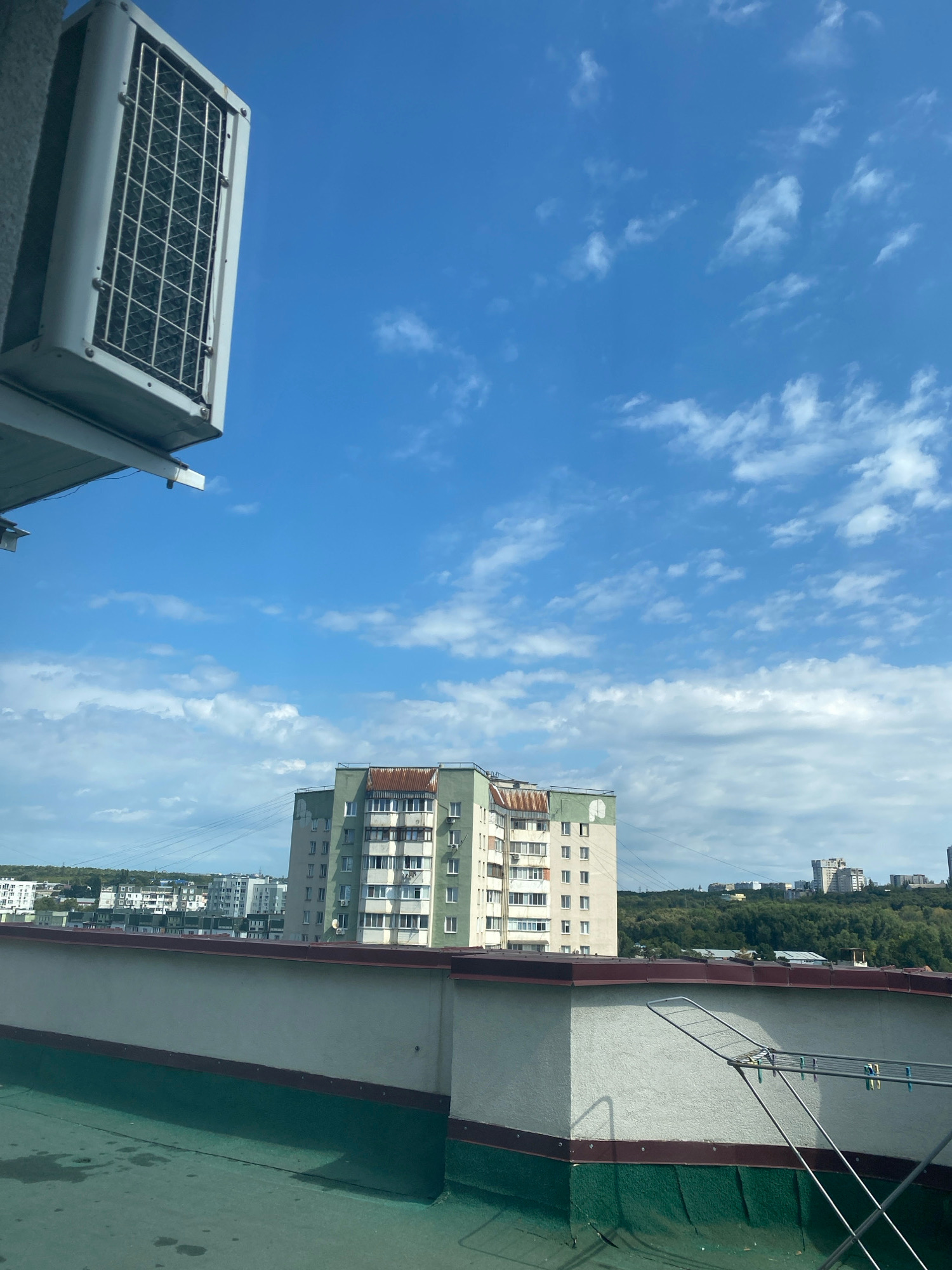 The image depicts a rooftop view in Chișinău, Moldova, under a bright blue sky with scattered white clouds. In the foreground, a rooftop surface is visible, with a textured green coating and a concrete parapet. An air conditioning unit is mounted on the left side of the frame, partially obscuring the sky. In the mid-ground, a multi-story residential building with a light green facade and a rusted metal roof is prominent. Several other buildings and trees are visible in the background, forming part of the urban landscape. A clothes drying rack is positioned on the parapet in the right foreground. No people are present in the scene.