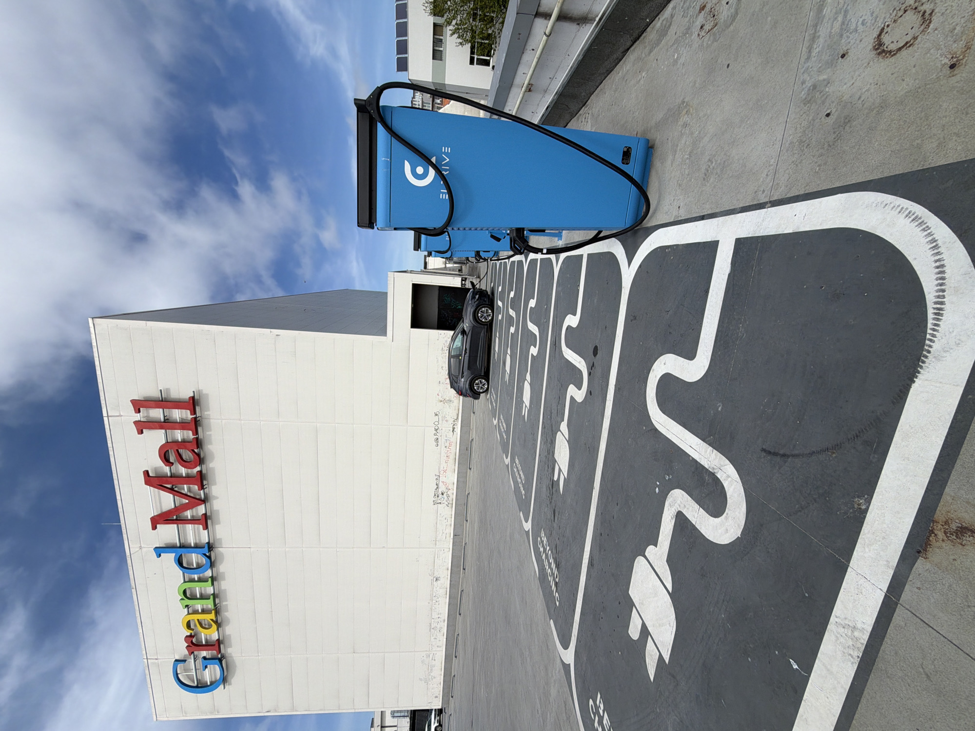 The image depicts an outdoor scene at the Grand Mall Varna in Varna, Bulgaria. A blue Electrify America EV charging station is prominently featured, with a black charging cable. Several parking spaces marked for electric vehicle charging are visible, each painted with a white outline and a plug graphic on a gray background. A gray car is parked in one of the spaces. The Grand Mall building is to the left with its name displayed vertically in red, blue, yellow, and green lettering. The sky is blue with wispy clouds. A gray concrete barrier and another building are also visible in the background.