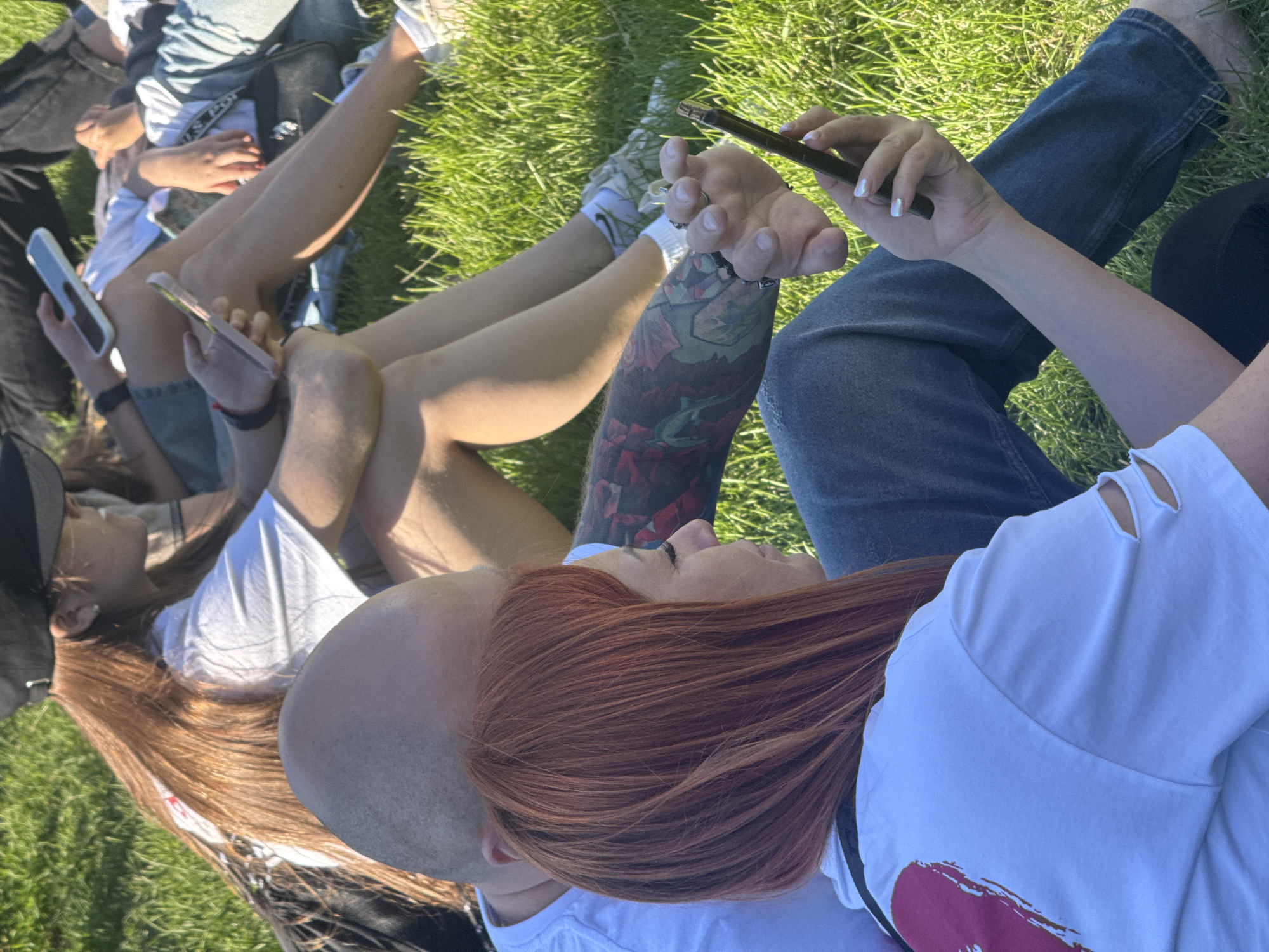 The image shows a group of people sitting on the grass, likely in a park, under bright sunlight, suggesting a warm, clear day. The scene is oriented vertically, with the subjects appearing to be lying down or reclining.
In the foreground, a person with striking red hair is lying down, looking up and to the right. They are wearing a white t-shirt with cut-out details on the shoulder. Next to them, and slightly behind, is the shaved head of another person. Their hands are intertwined, suggesting intimacy or close companionship. This person has a large, colorful tattoo covering their arm, featuring floral and possibly animal motifs. They are holding a dark-colored phone with their right hand, with a manicured fingernail visible.
Further back, other individuals are also visible, some with their legs stretched out. One person is holding a silver smartphone. Another person's legs are prominent, wearing white socks and sneakers. The grass is green and appears well-maintained.
The overall atmosphere is casual and relaxed, with people engrossed in their phones and enjoying each other's company outdoors. The time of day is likely afternoon, given the strong sunlight casting shadows and highlighting the textures of the grass and clothing. No visible text is present in the image.