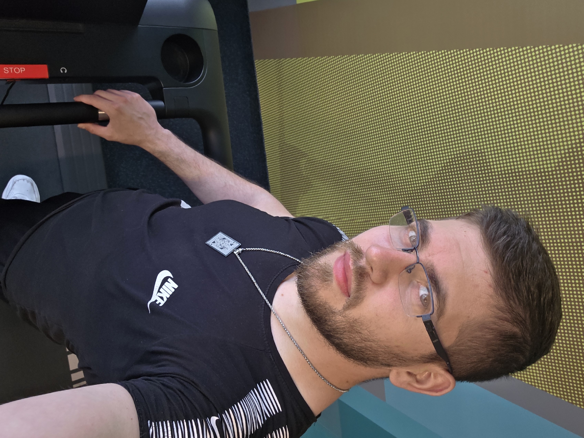 A man stands on a treadmill in a gym. He is wearing a black t-shirt with a Nike logo and a silver chain necklace. He is holding onto the treadmill's handlebars. He has a beard and is wearing glasses. The background is a yellow wall with a dotted pattern. The location is Plovdiv, Bulgaria.