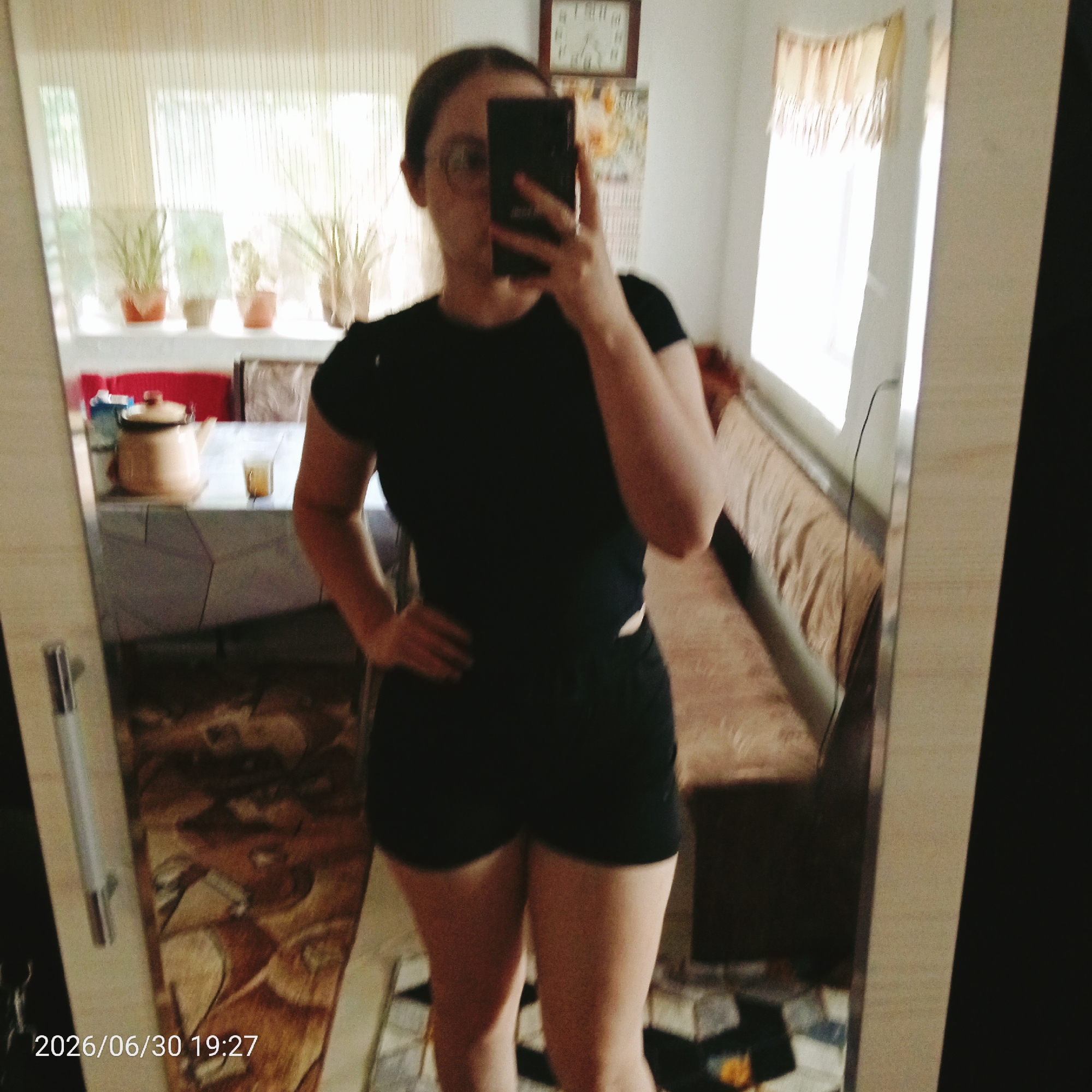 A person, partially visible, stands in front of a mirror, taking a selfie with a phone. The person is wearing a black t-shirt and black shorts. They have glasses on and their hair is pulled back. In the background, a window with sheer curtains allows diffused light into the room. Several potted plants are visible on the windowsill. A clock with a white face and Roman numerals hangs on the wall. The reflection in the mirror shows a table with a teapot and a glass, and further in the background, a couch. The flooring appears to be a patterned rug. The bottom left corner of the image displays the text "2026/06/30 19:27". The overall scene suggests an indoor setting, possibly a home, during the evening.