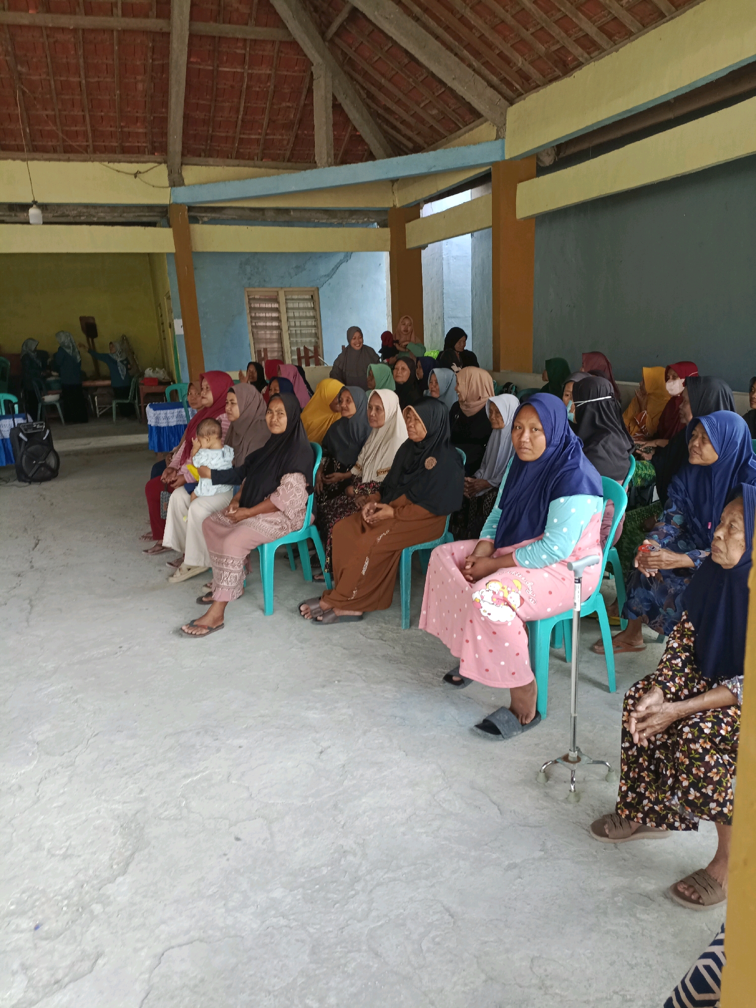 The image depicts an indoor gathering of approximately 20 individuals, predominantly women wearing headscarves, seated on chairs arranged in rows. The setting appears to be a community hall or a covered outdoor area with a tiled roof supported by wooden beams. The walls are painted in various colors, including yellow, blue, and a muted green. A speaker, a cane, and a few small tables with decorative cloths are visible. The floor is concrete. The individuals are mostly looking forward, suggesting they are attending an event or presentation. A baby is being held by one of the women. The overall atmosphere seems relaxed and communal. The location is Rancang Kencono, Indonesia.