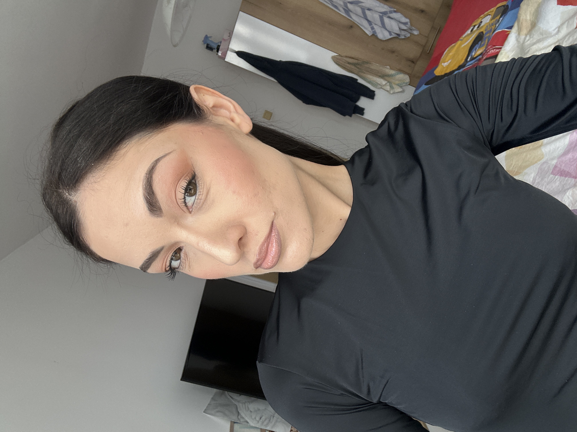 A medium-shot, selfie-style image shows a woman with fair skin, dark hair pulled back, and makeup including blush and lip color. She is wearing a black, long-sleeved top. The background includes a white wall, a TV mounted on the wall, a white cabinet with some items on top, including clothing, and a bed with cartoon-themed bedding. The exact location is not specified.