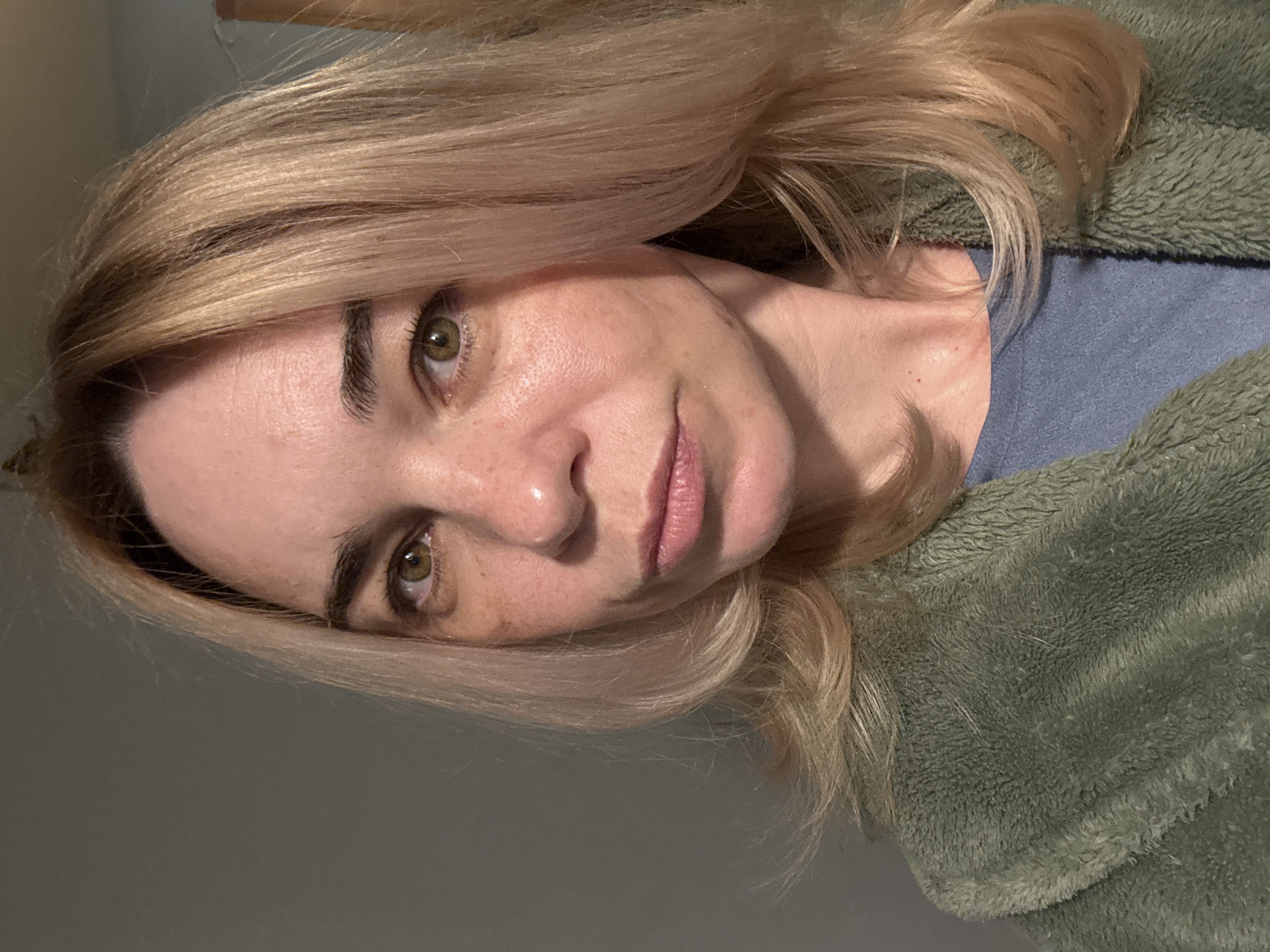 A woman with blonde hair and green eyes is facing the camera. She is wearing a blue shirt and a green fleece jacket. The background is a light gray wall. There is no indication that this image was taken in Vsevolozhsk, Russia.