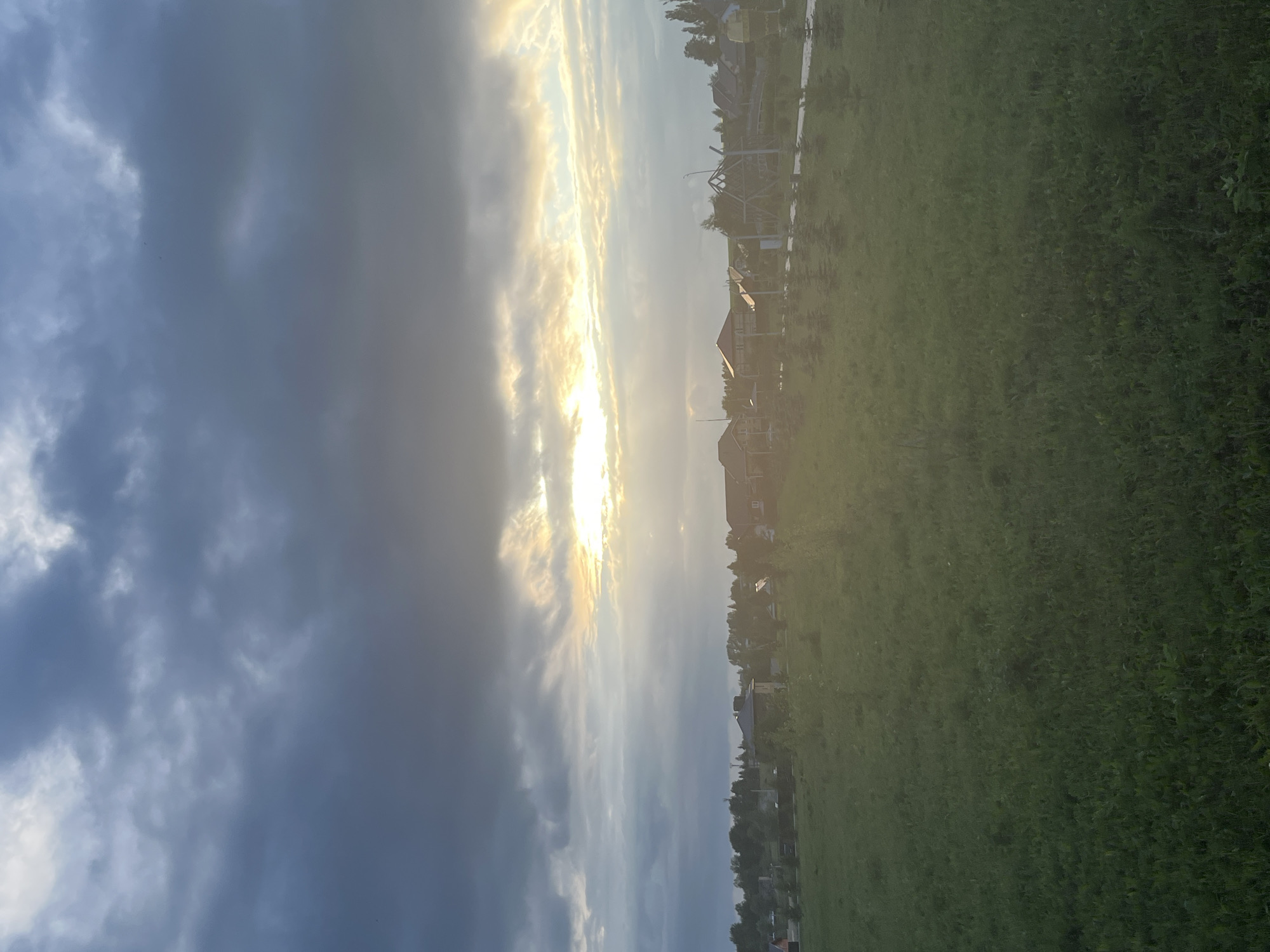 The image depicts a pastoral scene in Kamskiye Polyani, Russia, at dusk. The sky is dramatic, with dark, heavy clouds on the left giving way to streaks of golden light from the setting sun on the right. This transition suggests a clearing of a storm or approaching twilight. Below the sky, a line of houses with distinct roofs, indicative of rural or suburban architecture, is visible, nestled amongst trees. A partially constructed structure, possibly a building or framework, stands prominently near the houses. The foreground is dominated by a lush, green field, suggesting open land or a meadow. The overall impression is one of natural beauty, with the dynamic interplay of light and shadow creating a contemplative and serene atmosphere. The time of day is clearly evening, and the weather is transitioning, likely from rain to clear. There is no visible text in the image.