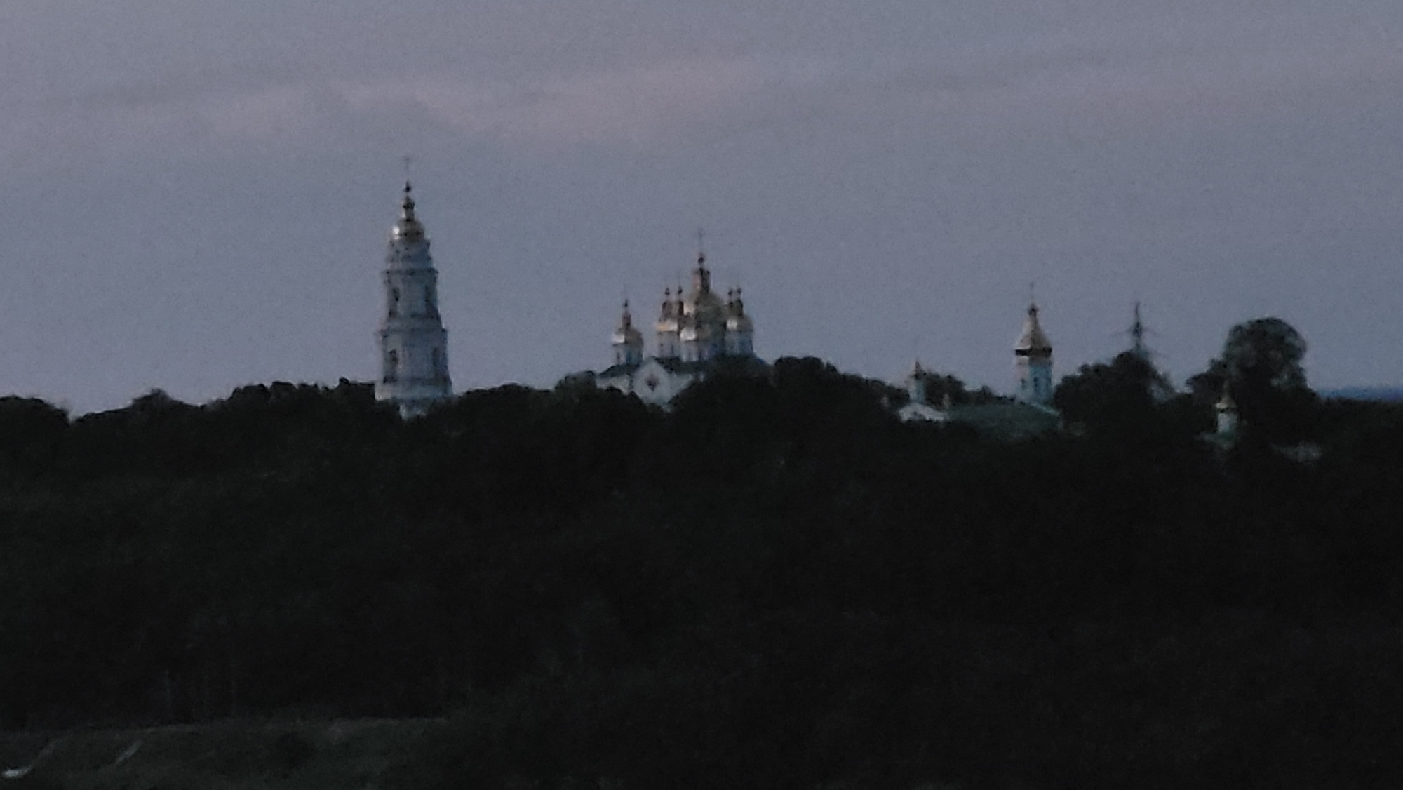 The image shows a silhouette of a monastery or church complex against a dusky sky. The main subjects are several buildings with domes and a tall bell tower, all appearing white or light-colored. These structures are situated on a hilltop, partially obscured by the dark silhouette of trees in the foreground. The sky is a uniform grey, suggesting either a cloudy day or twilight. The overall mood is serene and somewhat solemn due to the dim lighting and the nature of the structures depicted. The scene is likely a landscape view of a religious site. There is no visible text in the image.