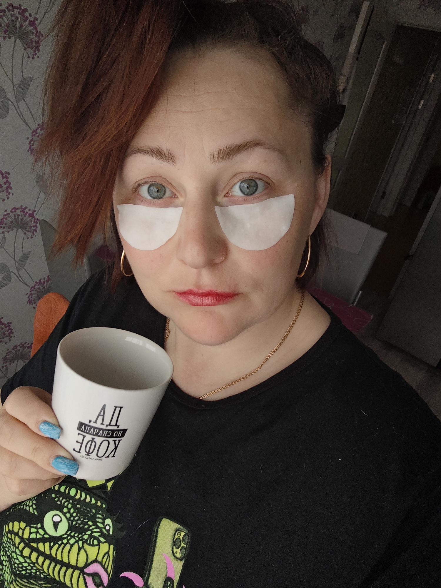 A woman with red hair and fair skin, wearing eye patches and red lipstick, is holding a white coffee mug with Cyrillic lettering ("АД АПАРЯНС ОН КОФЕ") in her left hand. She has blue painted nails with a wave design. She is wearing a black t-shirt with a lizard graphic and a thin gold necklace, along with gold hoop earrings. She is looking at the camera. The background includes a wall with floral wallpaper, a hallway, and a chair. The text on the mug translates to "AD Apperance On Coffee". The phone in her pocket has a light green case. Given the text on the coffee mug, it's likely that the location is Markovskii, Russia.