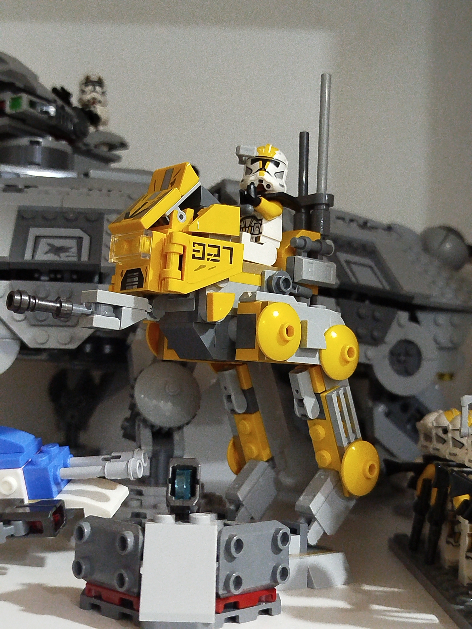 The image displays a collection of LEGO Star Wars models. In the foreground, a yellow and gray AT-RT walker with a Clone Trooper pilot is positioned. The walker has a detailed construction with visible studs and hinges. Behind it, a large gray AT-TE walker is partially visible. To the left of the AT-RT, a blue and white LEGO construction is present. To the right, the legs of several LEGO Clone Troopers are visible. The background is a plain, light-colored surface, likely a shelf. The lighting suggests an indoor environment. There is no information available regarding the location in Mirny, Russia, as this is a fictional scene created with toys.