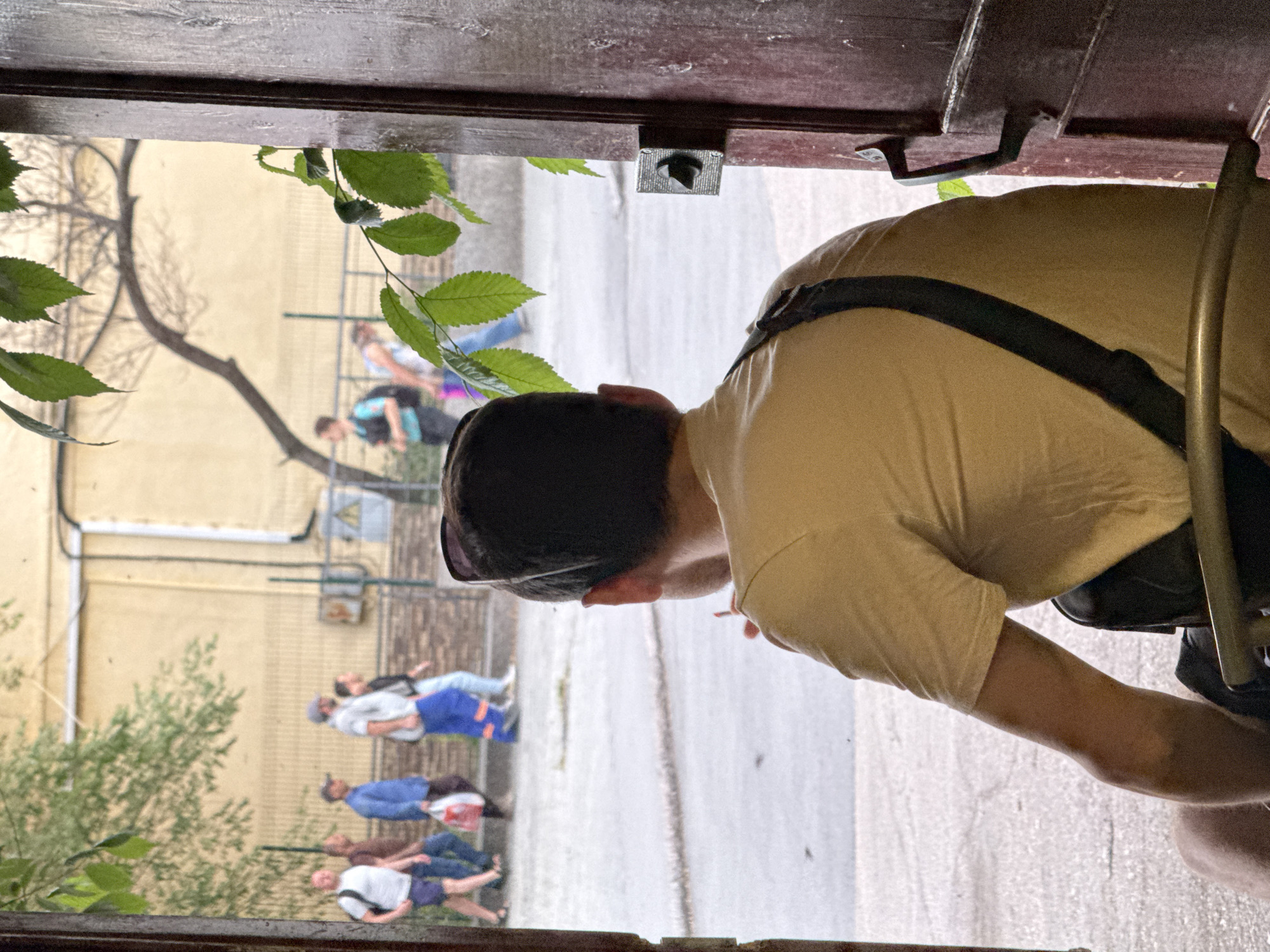 In the city of Unknown, Unknown, a man sits facing away from the camera, looking out an open doorway. He is wearing a light-colored t-shirt and sunglasses are pushed up on his head. He appears to be holding a cigarette in his right hand. Behind him, the doorway reveals a street scene with several people walking. The building behind the pedestrians is a light yellow color, with a dark wooden fence in front of it. Green leaves are visible in the foreground, obscuring part of the scene. The overall impression is of a casual, outdoor environment, possibly a cafe or a residential street.