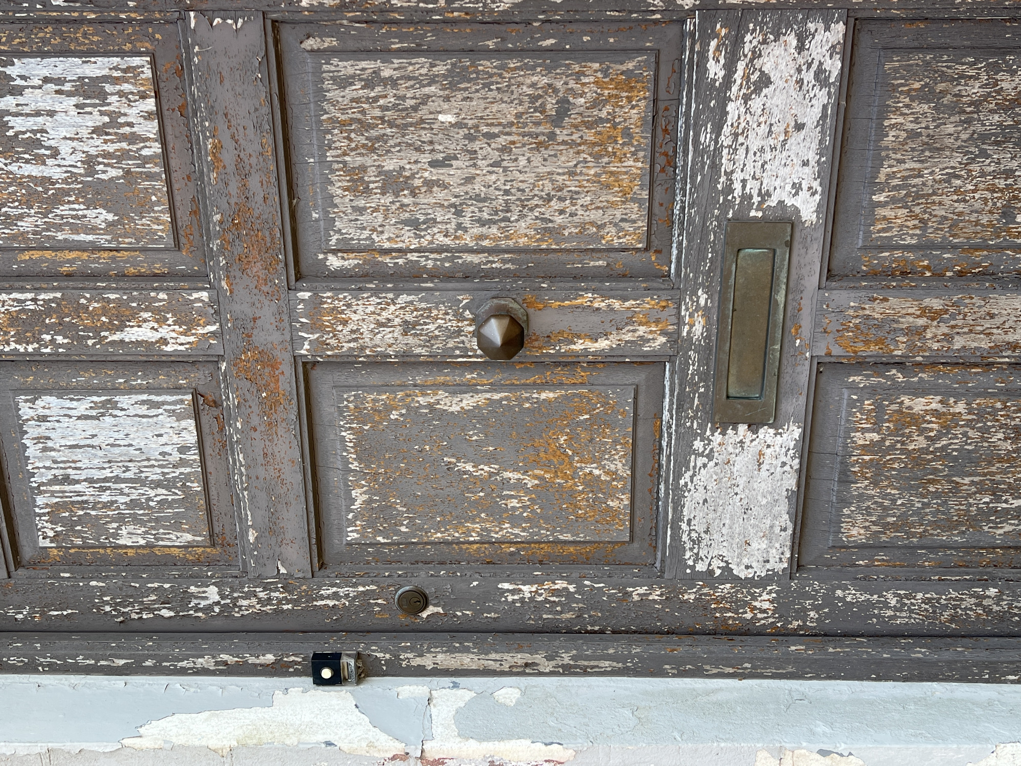 The image shows a close-up of a weathered, old wooden door with peeling grey and white paint, revealing a reddish-brown undercoat. The door is divided into multiple panels. Visible hardware includes a round doorknob, a keyhole, a letterbox slot, and a small black and silver intercom unit mounted to the adjacent wall. The wall itself is painted a light, uneven blue-grey color and shows signs of damage and peeling paint. The scene is presented in a static, unposed manner, suggesting an outdoor urban setting. The specific location is a door on a building in the town of Mountcharles, County Donegal, Ireland.