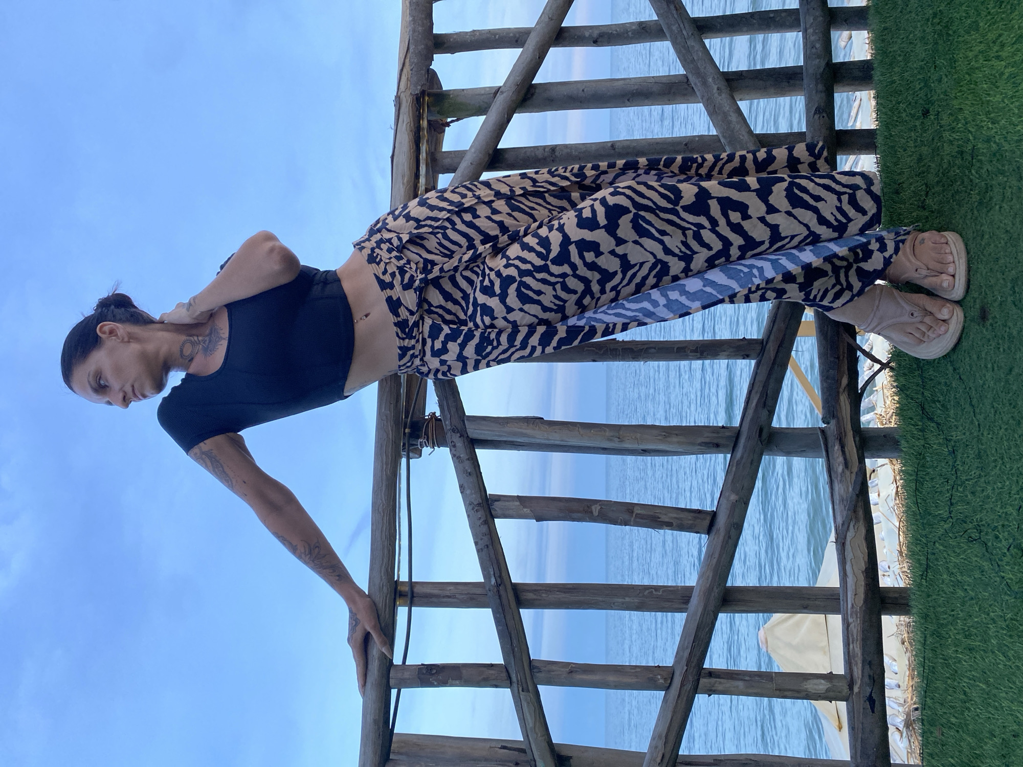 A woman with dark hair pulled back and tattoos on her arms and neck leans on a wooden railing against a blue sky and ocean. She wears a black crop top and a long, flowing skirt with a tan and black animal print. Her feet are adorned with beige sandals. The railing is constructed from natural, weathered wood and supports a pathway. In the background, the blue ocean stretches to the horizon, with a hint of white structures and green grass in the foreground. The image appears to be taken outdoors, possibly at a beachside location. The specific location in Phu Thuy Ward, Vietnam is not evident from the visual details.