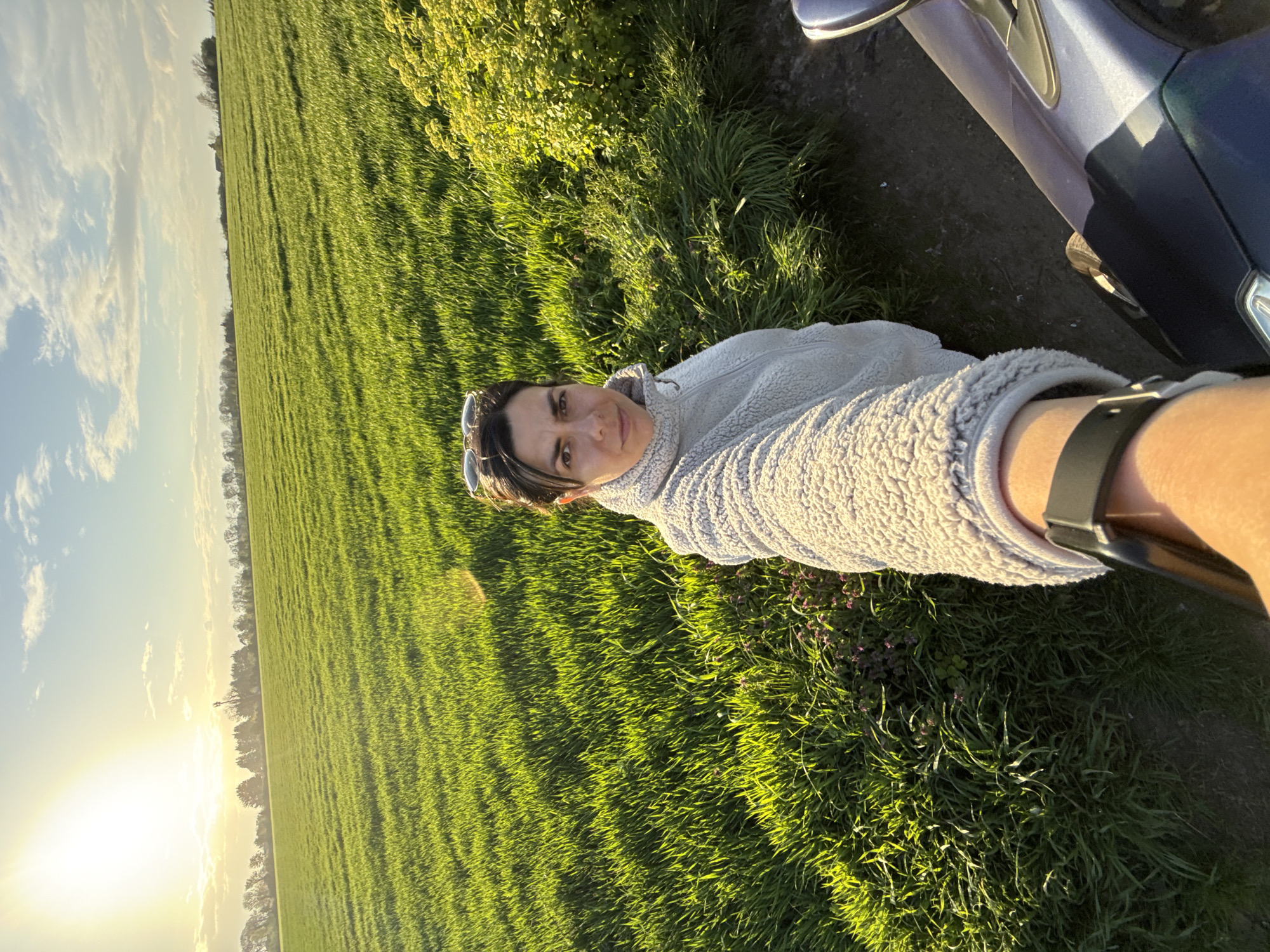 A woman is standing near a car next to a grassy field at sunset. The woman is wearing a light-colored fuzzy sweater and a watch. Sunglasses are perched on her head. The car is blue. The field is green and stretches into the distance, with trees visible on the horizon. The sun is low in the sky. The location is near Great Yarmouth, United Kingdom.