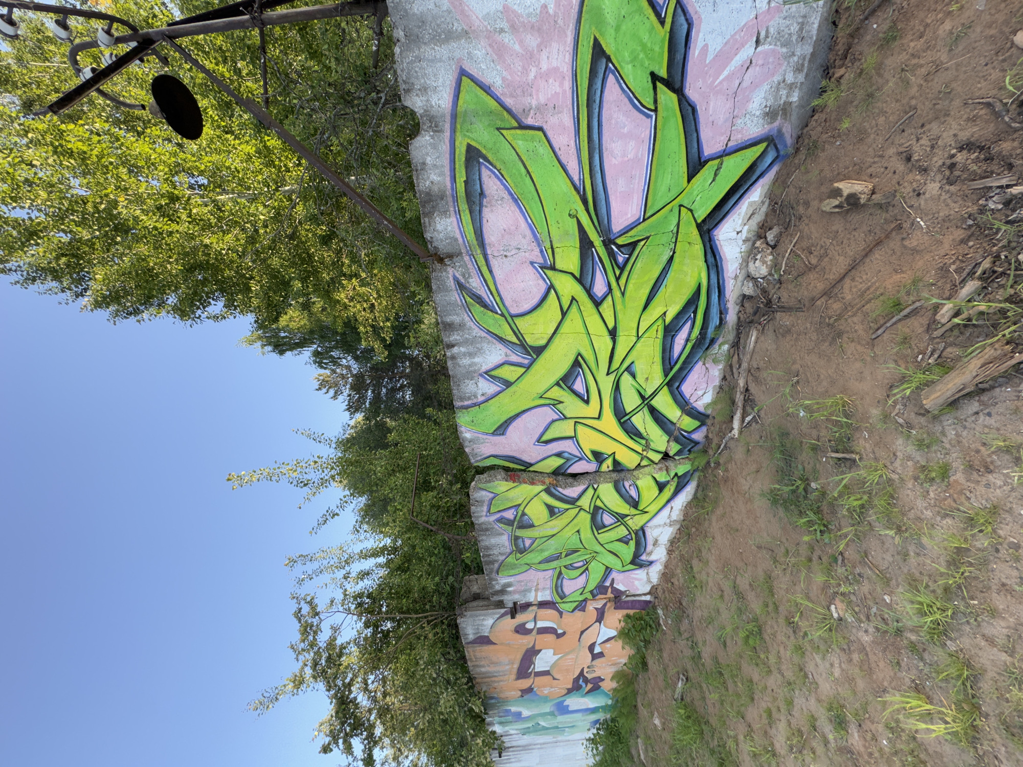 The image shows a concrete wall covered in graffiti. The largest piece of graffiti is predominantly green with blue and yellow accents and a pink background, appearing abstract. Below it, there's another graffiti piece with orange and brown hues. The wall appears cracked and damaged. Adjacent to the wall is a patch of dirt and grass. Trees and blue sky are visible behind and to the side of the wall. A metal pole with a black disc attached is visible above the trees. Based on the query parameters, the location is assumed to be in the city of Chaikovsky, Russia.